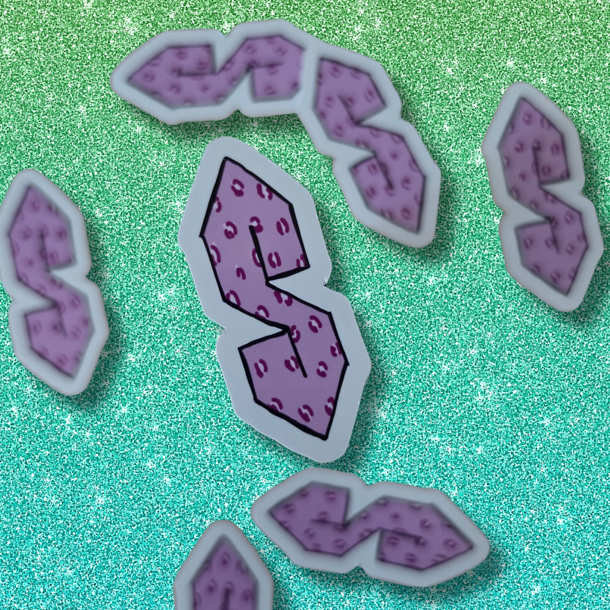 A set of purple and pink textured letter 'S' stickers with white borders, arranged on a sparkling green background.