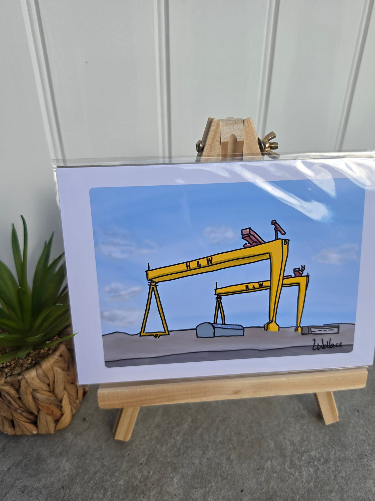 A framed art print depicting a yellow gantry crane on a blue sky background, displayed on a small wooden easel next to a potted succulent.