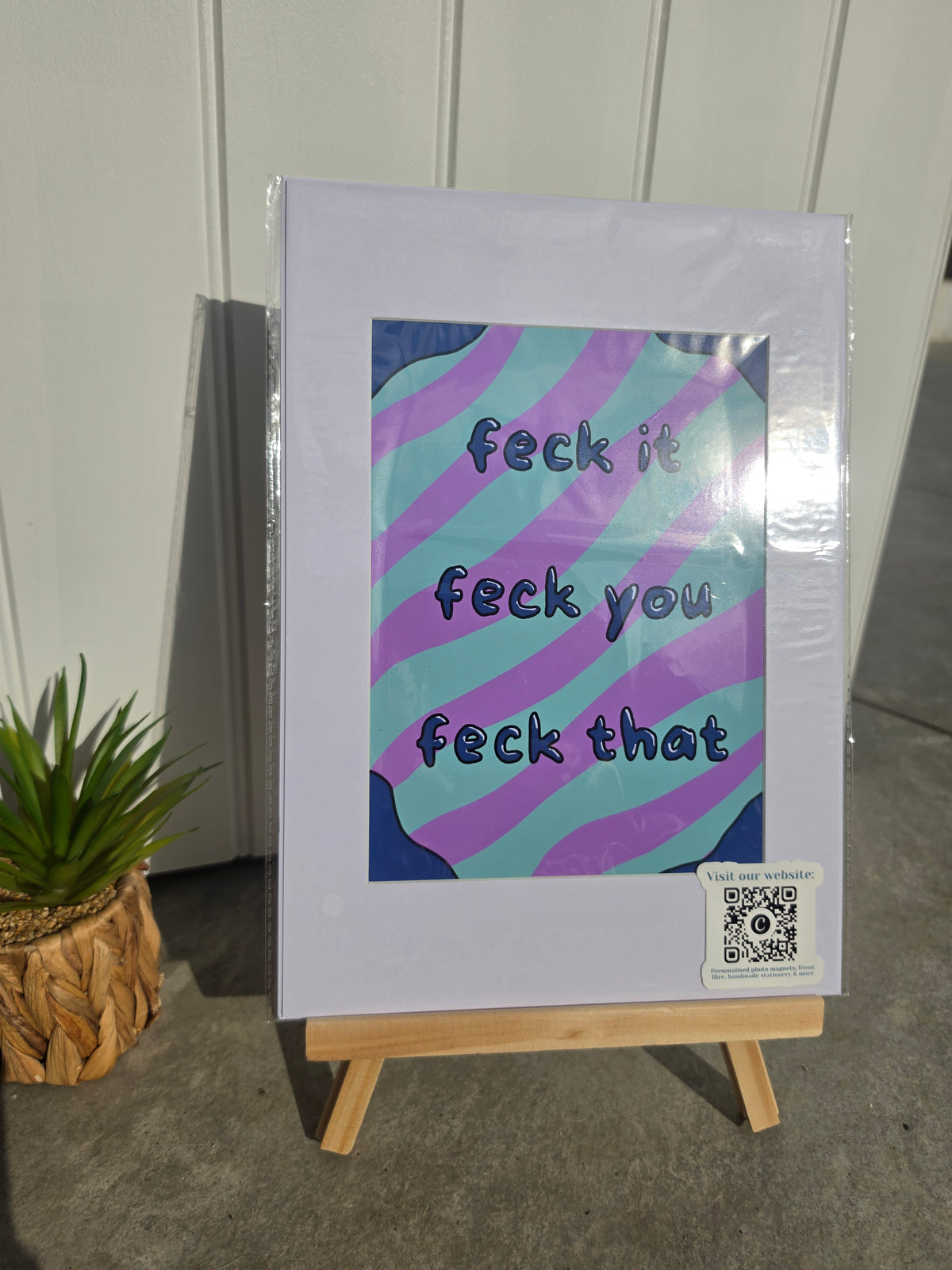 A printed art poster or sign displayed on a small wooden easel, featuring the phrase 'feck it feck you feck that' in dark lettering over wavy purple and teal stripes. The print is photographed against a white paneled wall with a small potted plant at the side.