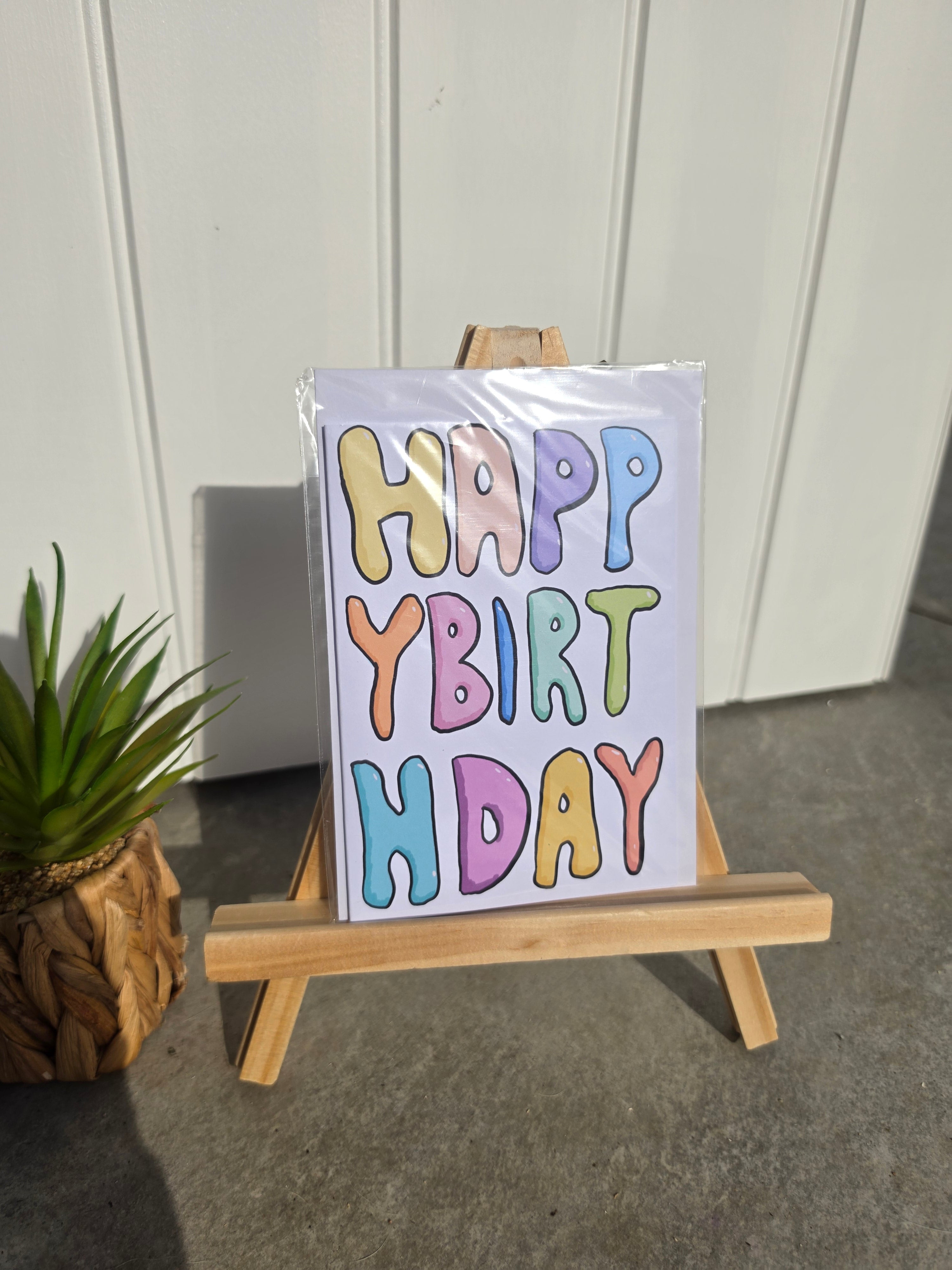 A birthday card with the words 'HAPPY BIRTH DAY' in colorful bubble letters, displayed on a small wooden easel. The card is wrapped in clear plastic and set against a white paneled wall with a concrete floor, next to a potted green plant.