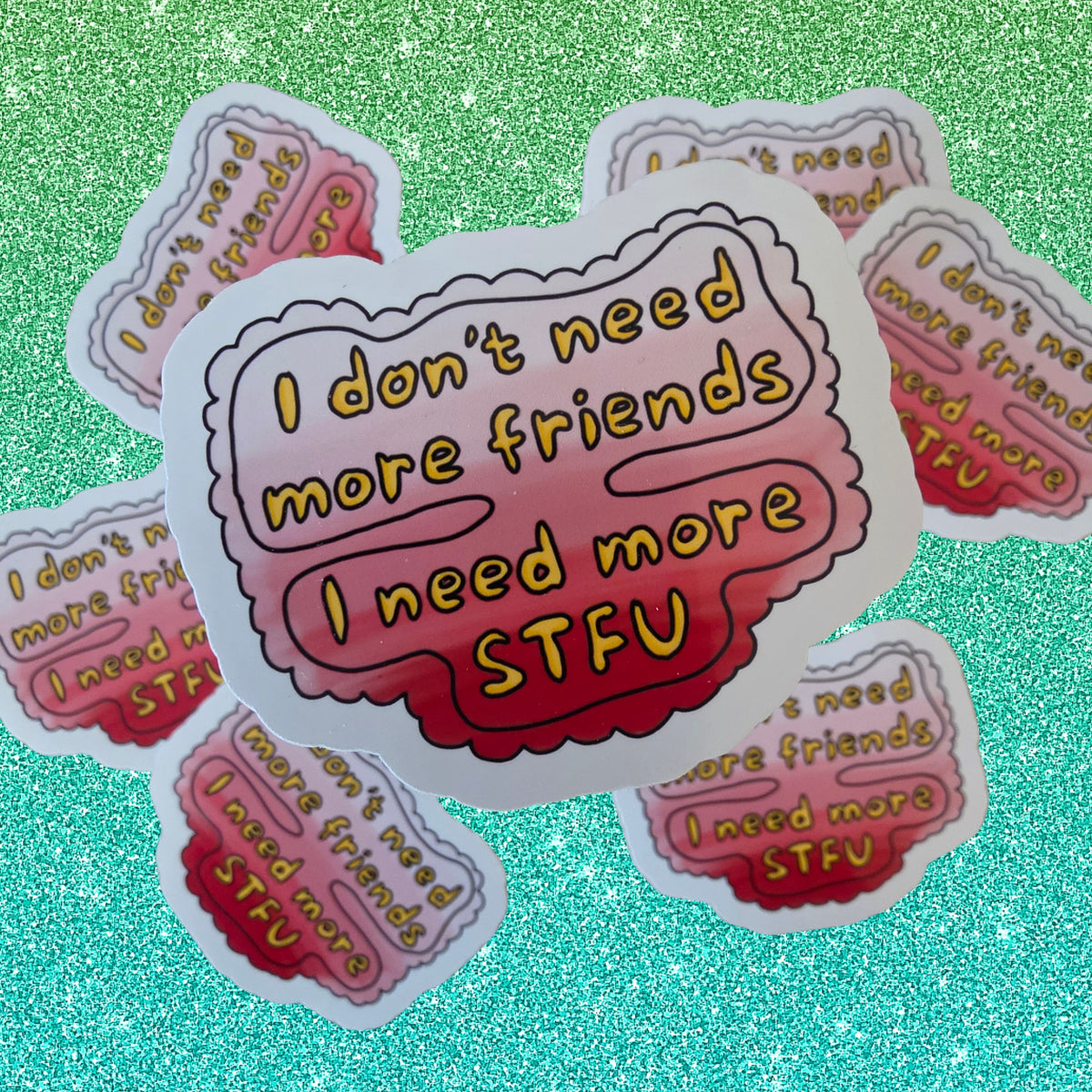A die-cut sticker with the text 'I don't need more friends I need more STFU' in yellow lettering over a pink-to-red gradient background, shown on a sparkling green surface with multiple identical stickers scattered around.