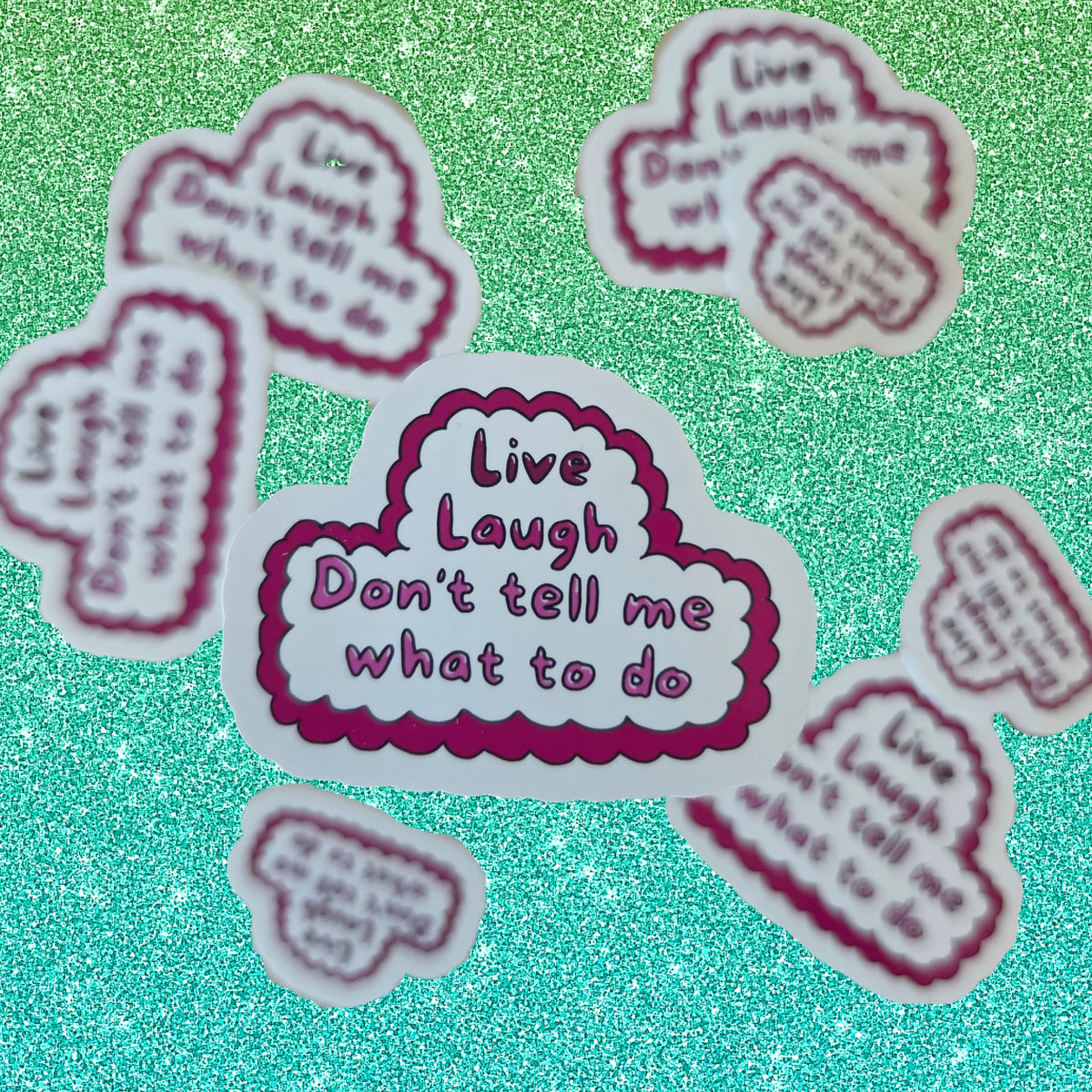 A set of die-cut cloud-shaped stickers with the text 'Live Laugh Don't tell me what to do' in purple lettering, featuring a pink wavy border and white background, arranged on a sparkling teal surface.