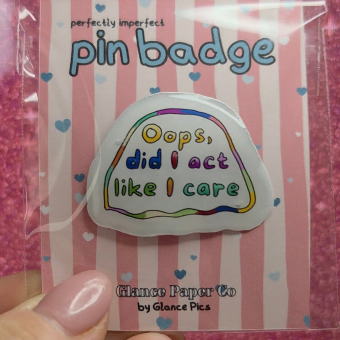 A hand holds a packaged RESIN pin badge against a pink glittery background. The pin badge is shaped like a cloud with the text 'Oops, did I act like I care' in rainbow-colored lettering.