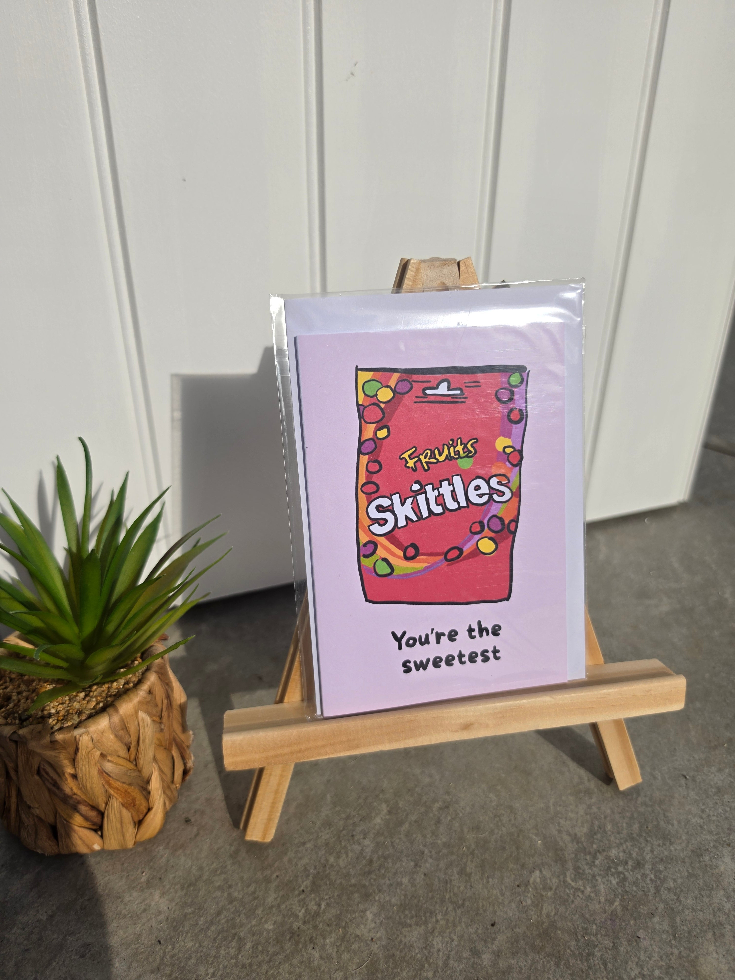 A greeting card displayed on a small wooden easel, featuring a pink background with a Skittles box illustration and the text 'You're the sweetest'. A small potted succulent sits to the left against a white paneled wall.