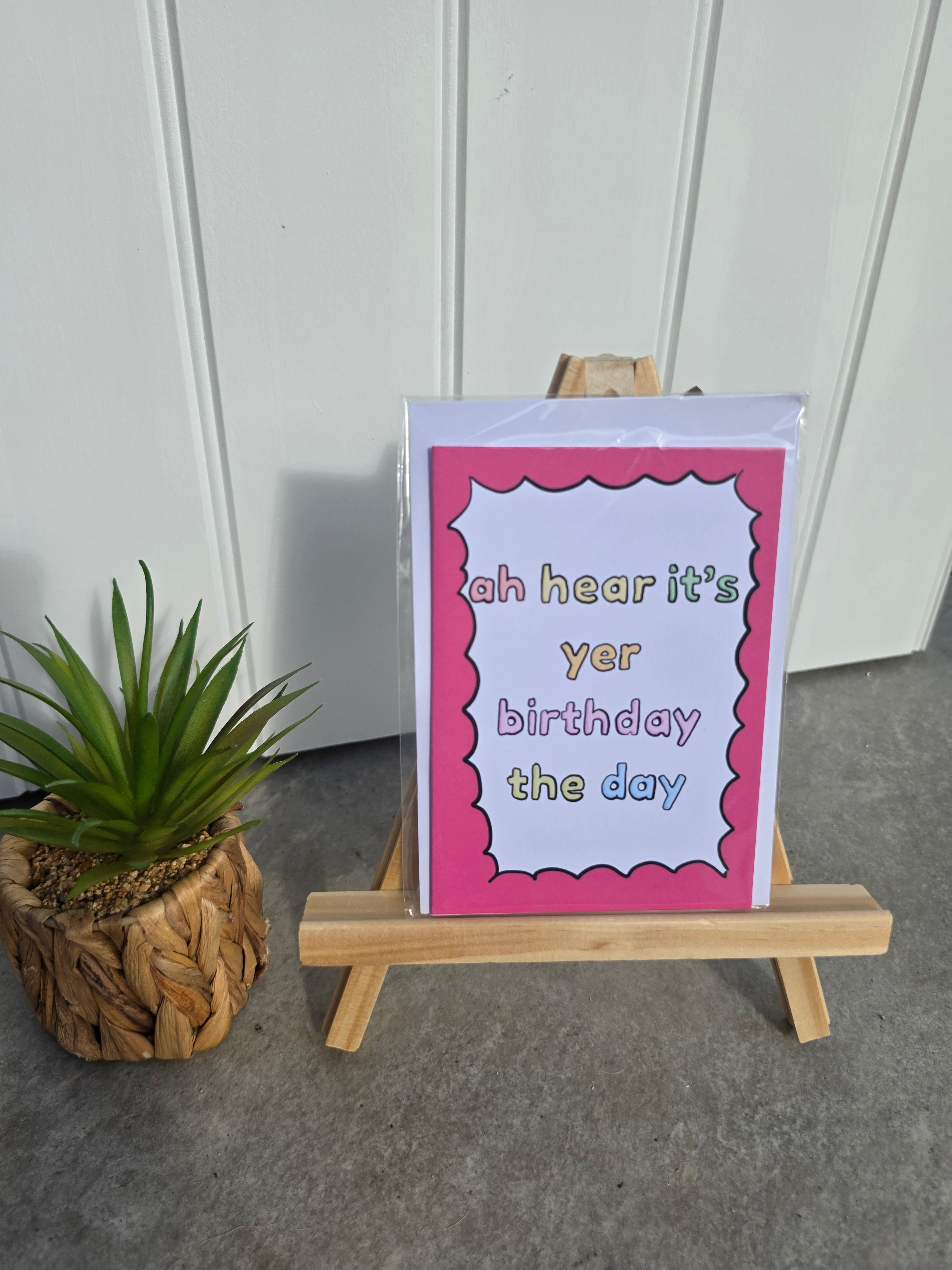 A greeting card with a pink border and playful text reading 'ah hear it's yer birthday the day' displayed on a small wooden easel, with a potted plant to the left against a white paneled wall.