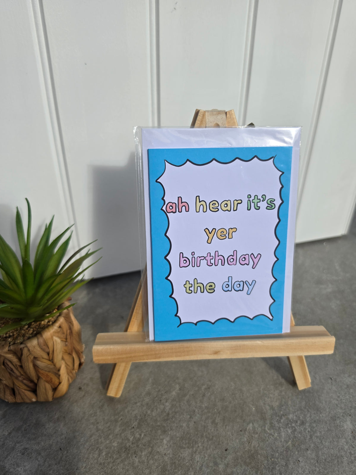 A greeting card displayed on a small wooden easel, with a potted plant to the left and a white paneled wall in the background. The card has a bright blue border and a white center with the text 'ah hear it's yer birthday the day' in multicolored lowercase letters.