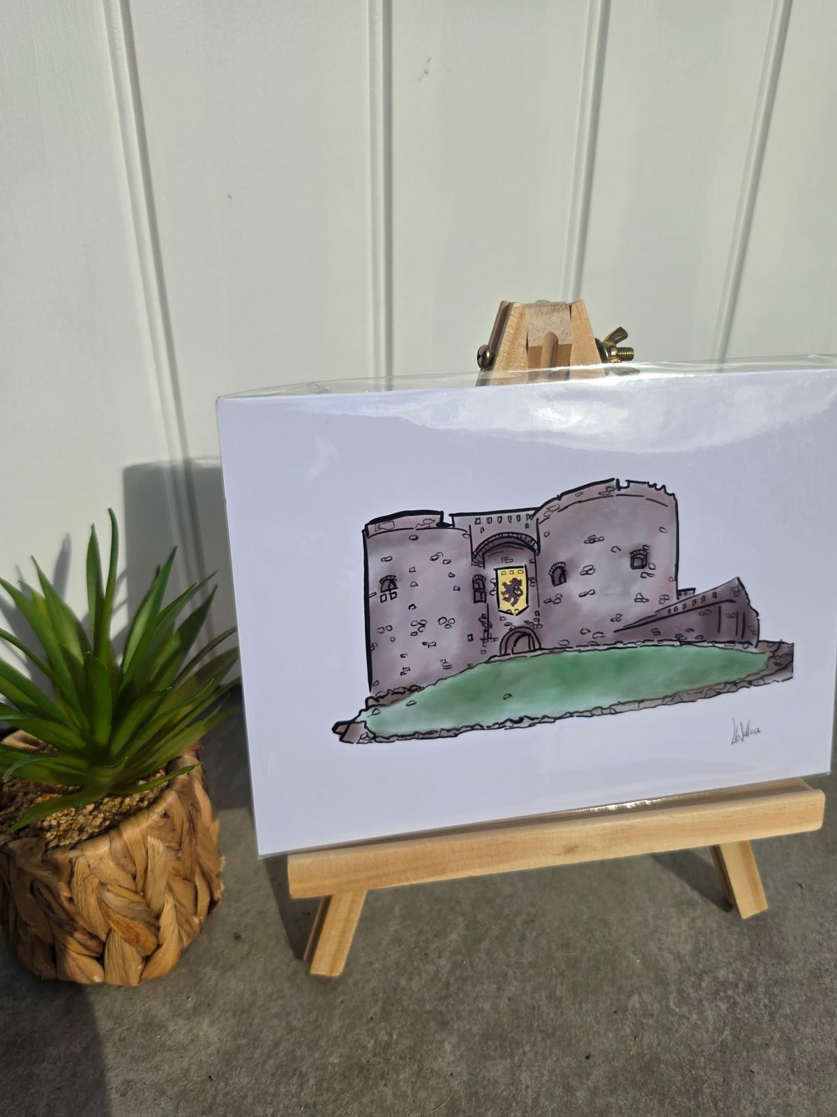 A printed art piece showing a hand-drawn castle with a green foreground and yellow shield, displayed on a small wooden easel. 