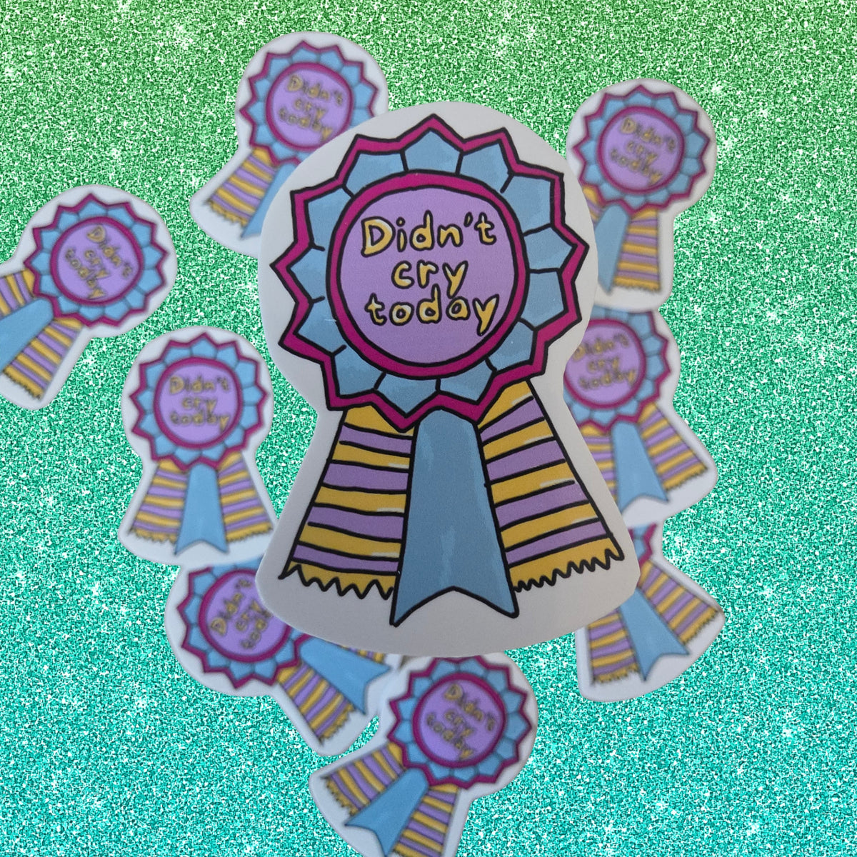 A set of hand-drawn style ribbon award stickers with the text 'Didn't cry today' in yellow on a purple center, surrounded by blue and pink stripes, arranged on a glittery teal background.