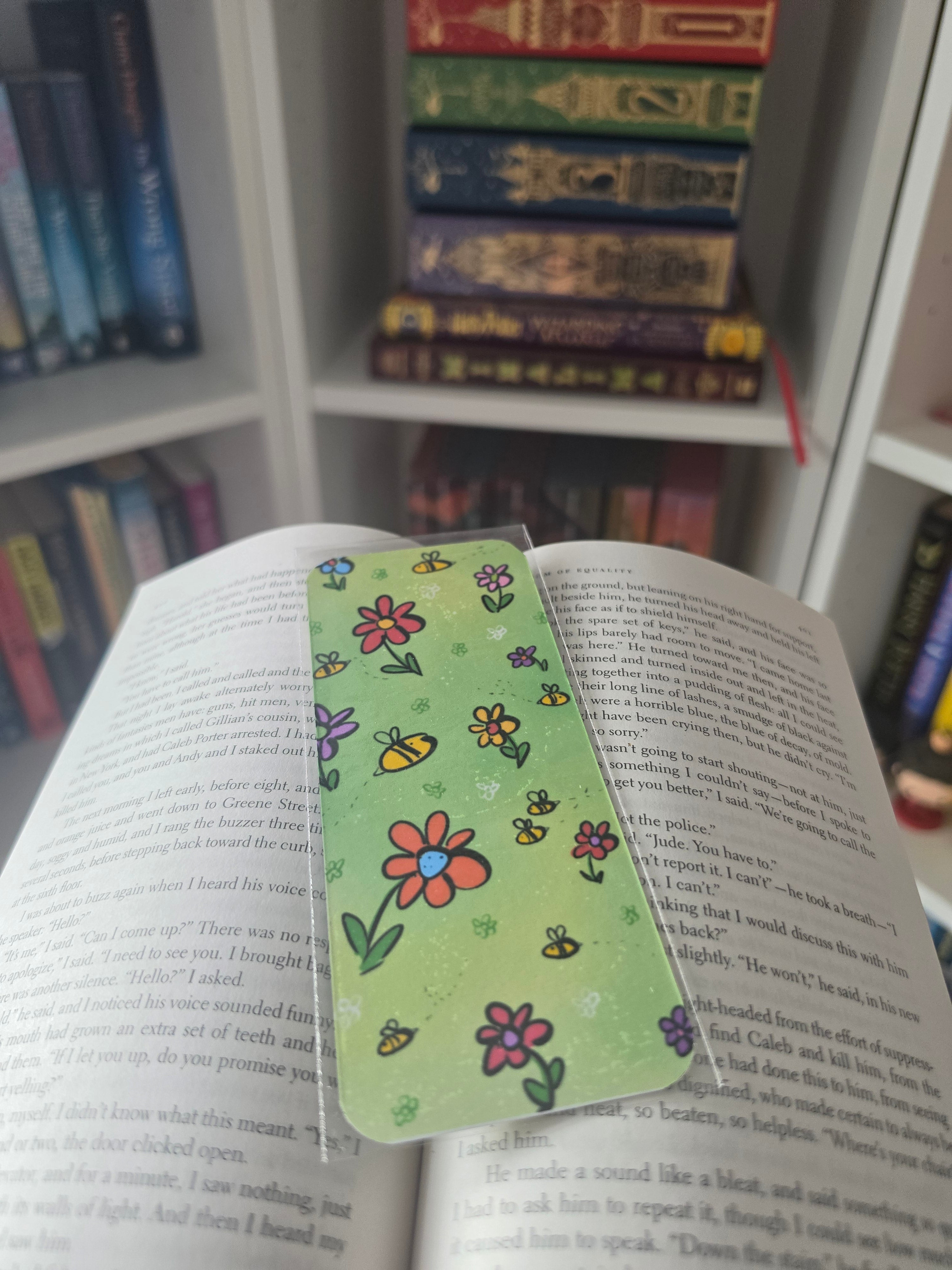 A clear plastic book bookmark with a light green background and colorful hand-drawn flowers and bees, shown resting on an open book in front of a blurred background of bookshelves.