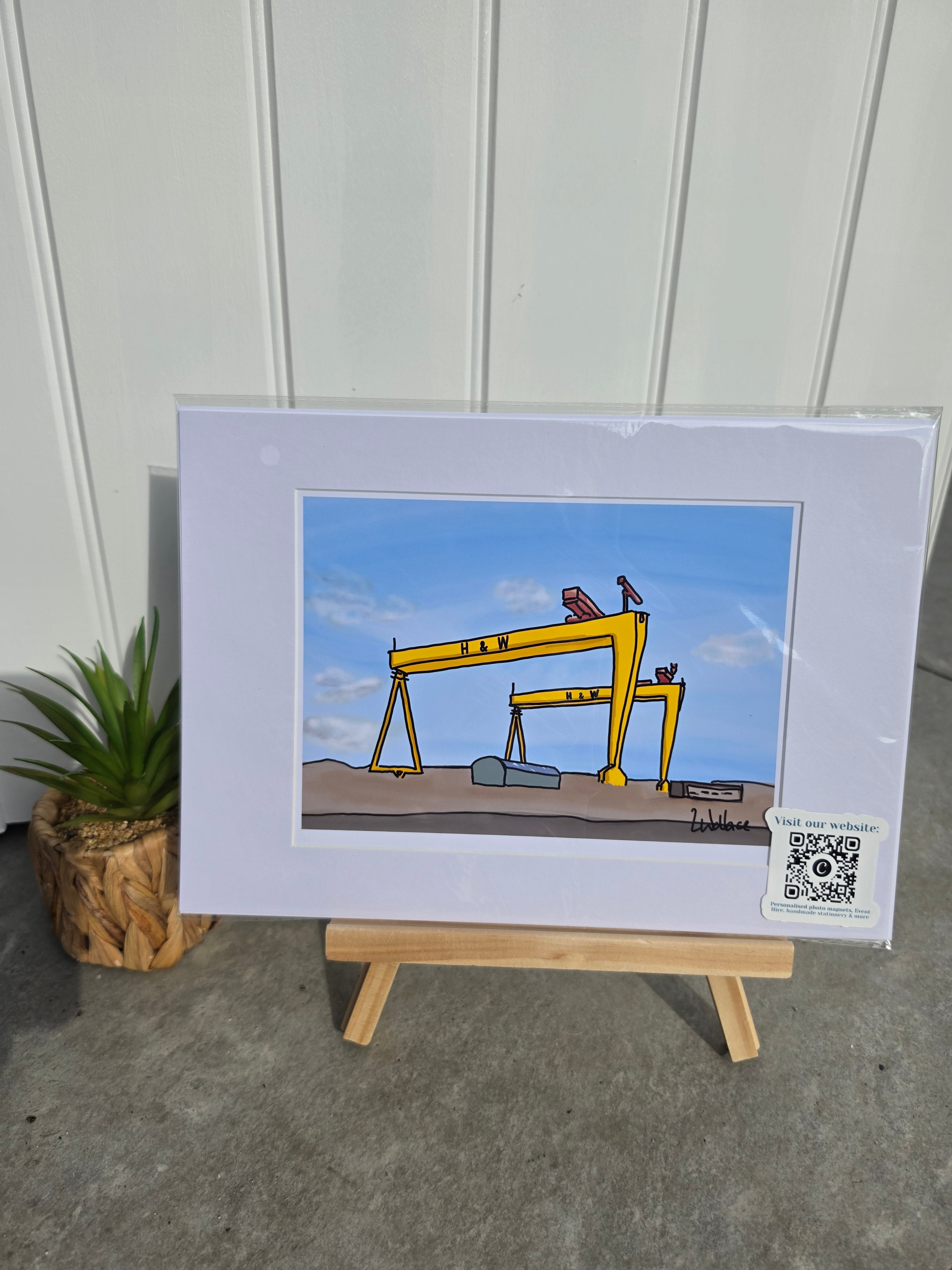 A matted art print displayed on a small wooden easel against a white paneled wall, featuring a cartoon illustration of yellow cranes under a blue sky. A small plant in a wood planter sits to the left, 