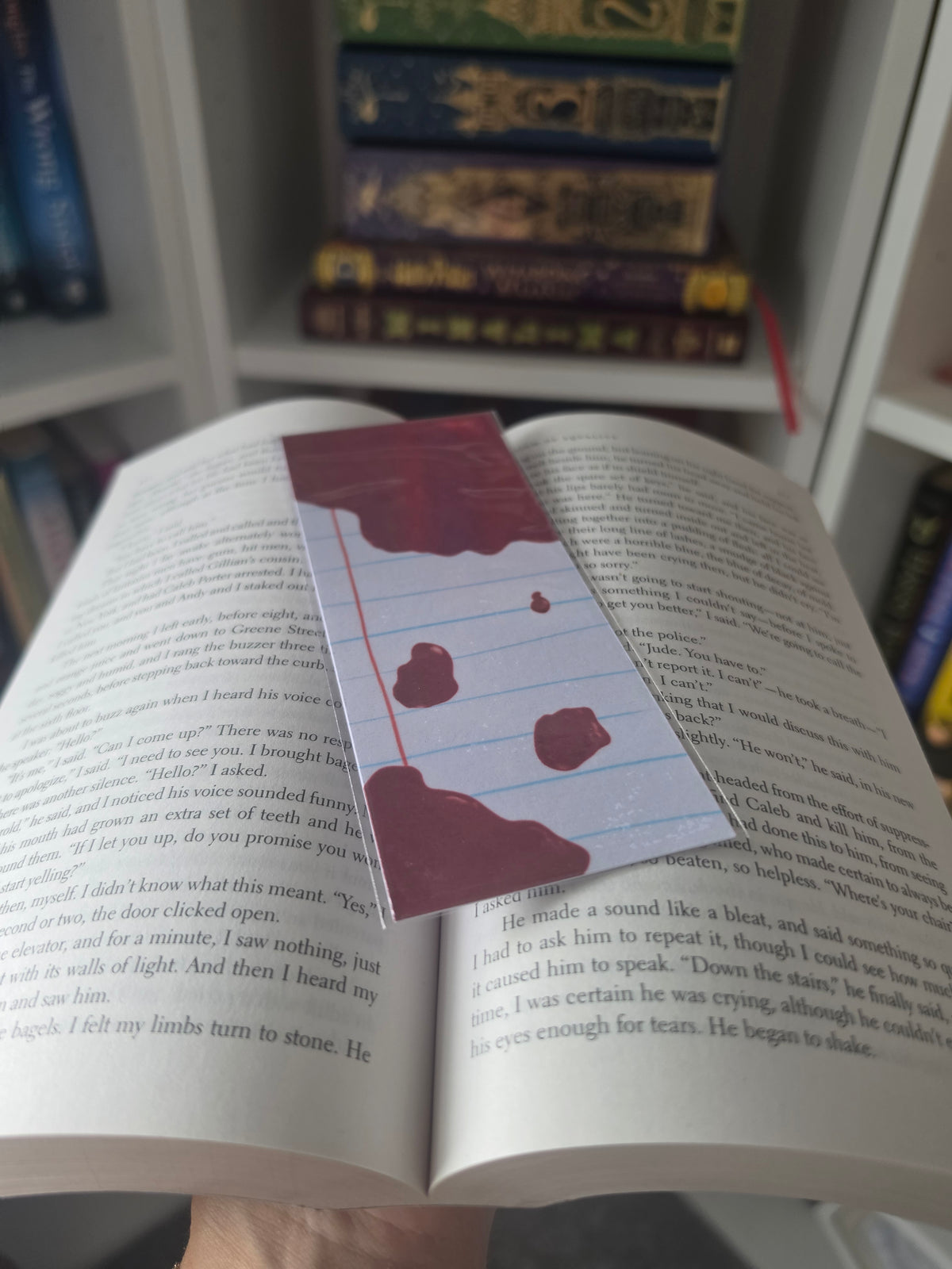 A close-up of an open book with printed text, featuring a rectangular bookmark or card with lined paper design and dark red blood-like splatters, held in a hand against a background of stacked books.