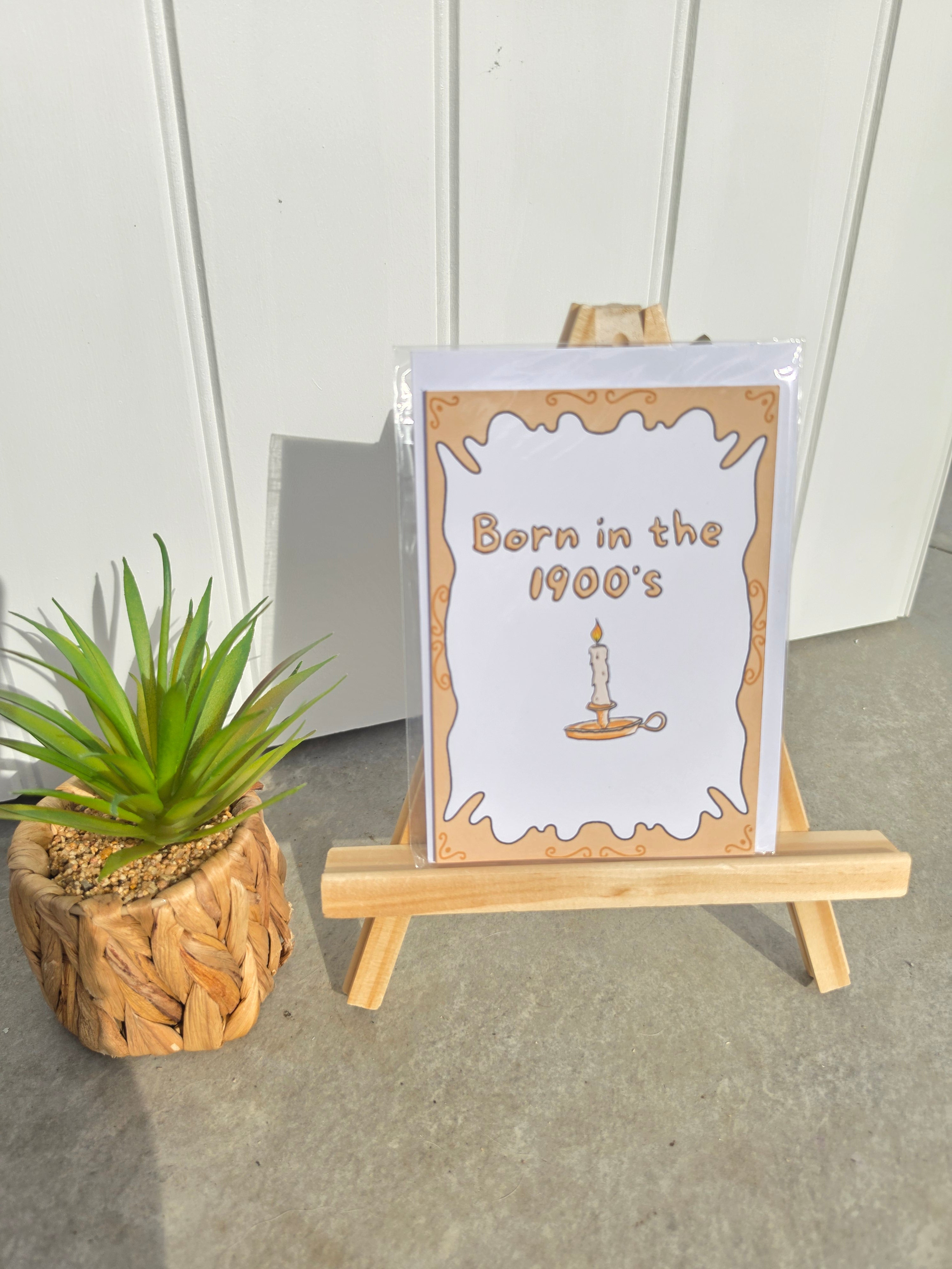 A greeting card that reads 'Born in the 1900's' with a candle illustration, displayed on a small wooden easel next to a potted succulent.