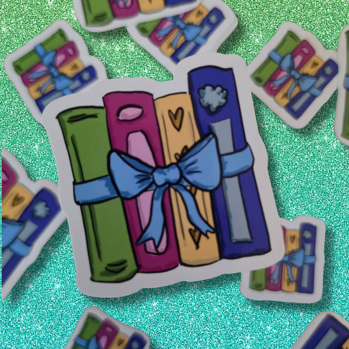 A die-cut sticker featuring a cartoon illustration of colorful books tied with a blue ribbon and bow, displayed on a sparkling teal background with other similar stickers scattered around.