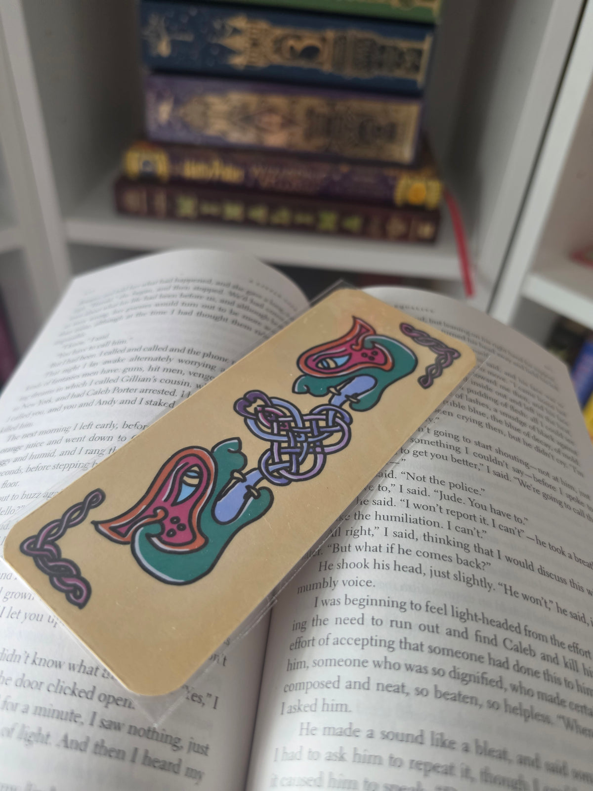 A rectangular bookmark with colorful Celtic knot illustrations rests on open book pages, with stacked books blurred in the background.