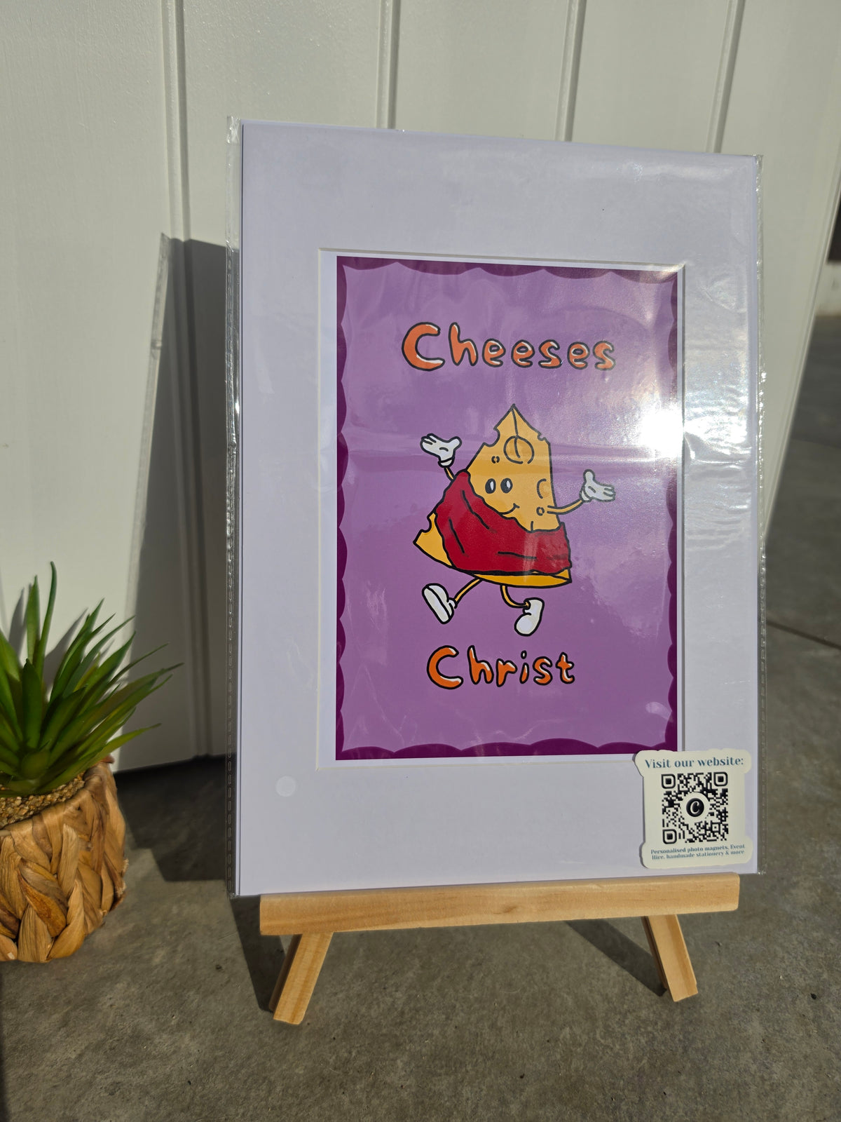 An art print displayed on a small wooden easel. The print features a purple background with a cartoon illustration of a cheese character wearing a red robe and the words 'Cheeses Christ' in orange.