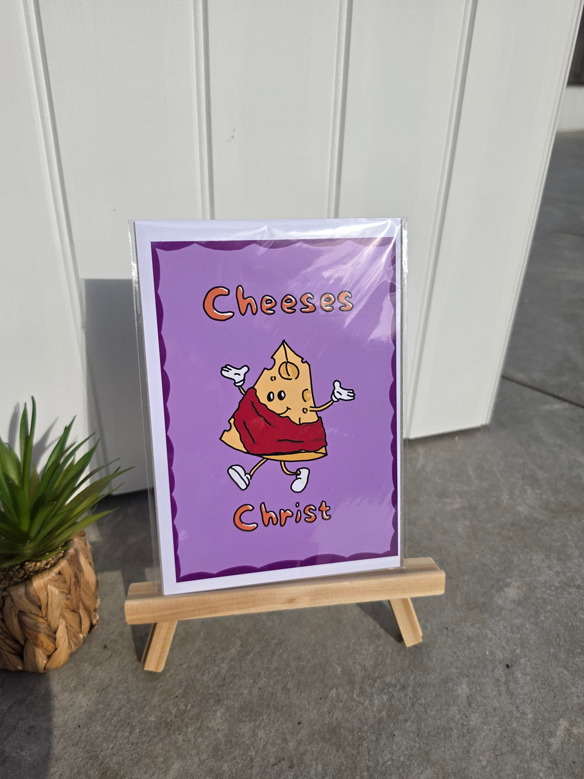 A custom printed art card featuring a cartoon cheese slice character with the text 'Cheeses Christ' on a purple background, displayed on a small wooden easel outdoors.