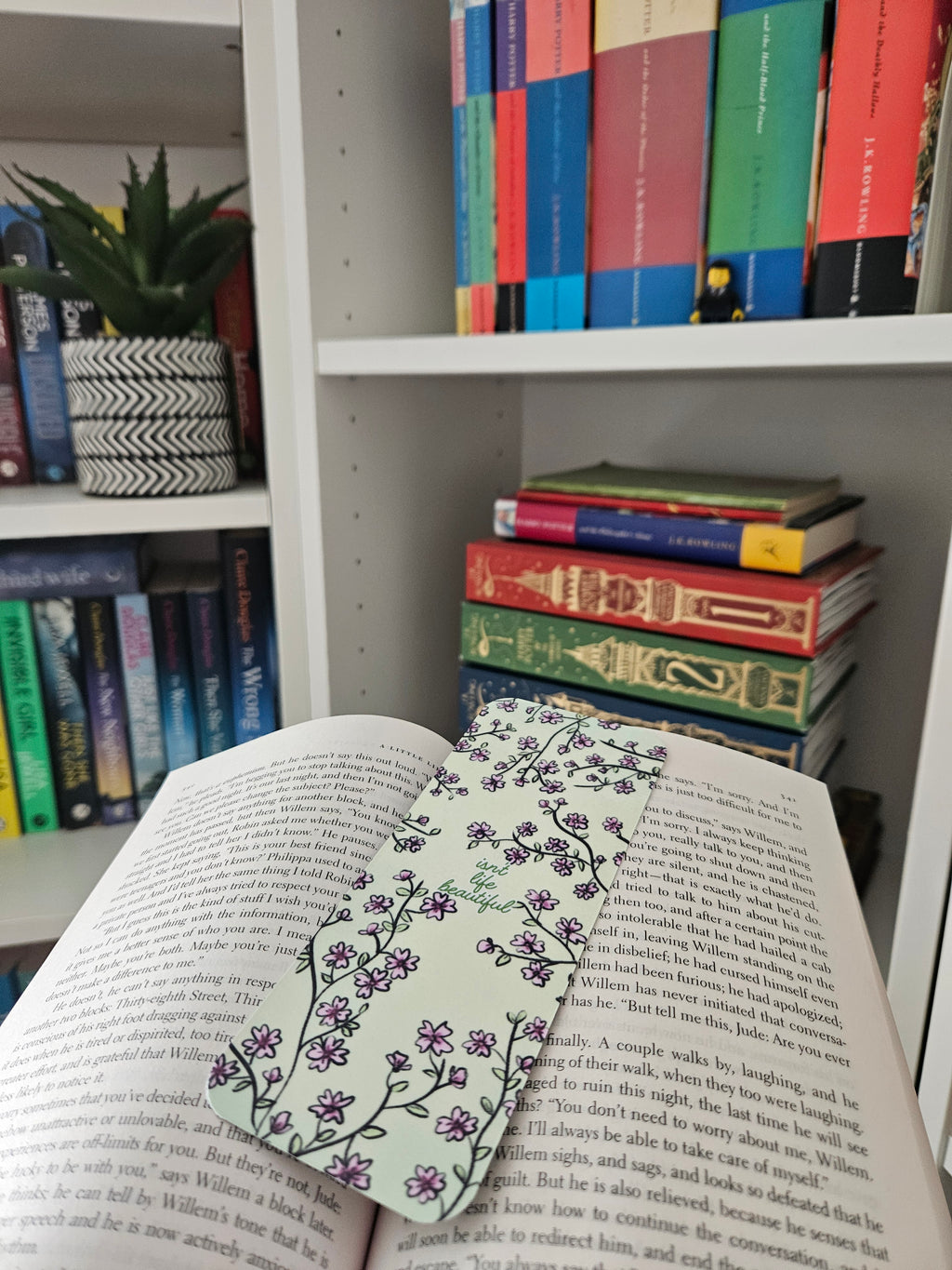 An open book in front of a white bookshelf filled with books. A decorative bookmark with a pink floral vine pattern is draped across the pages.