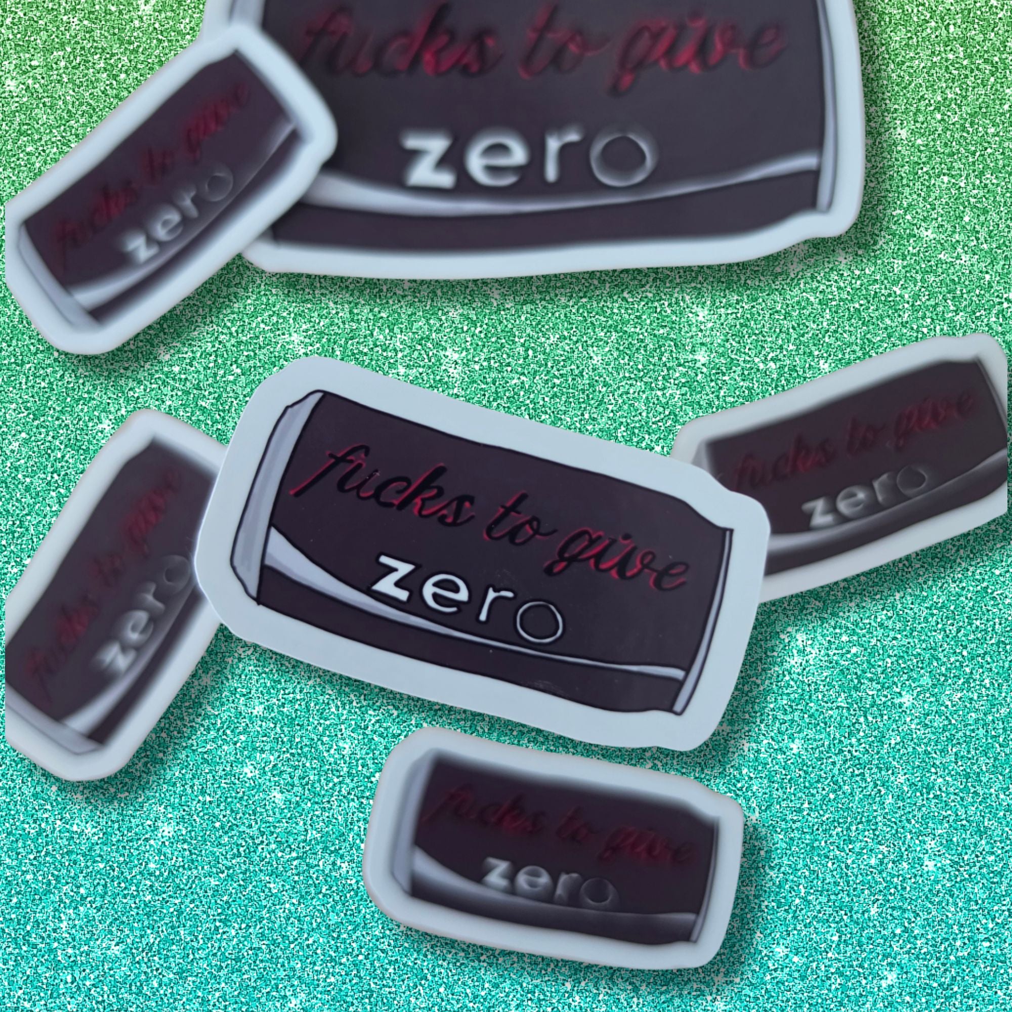 Several small drinks can stickers scattered on a glittery green background with the phrase "fucks to give zero" in red script and white text.