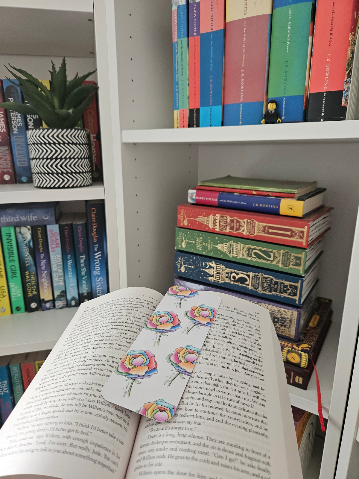 A white bookmark with colorful watercolor-style peonies rests on an open book on a white bookshelf, with stacks of books in the background.