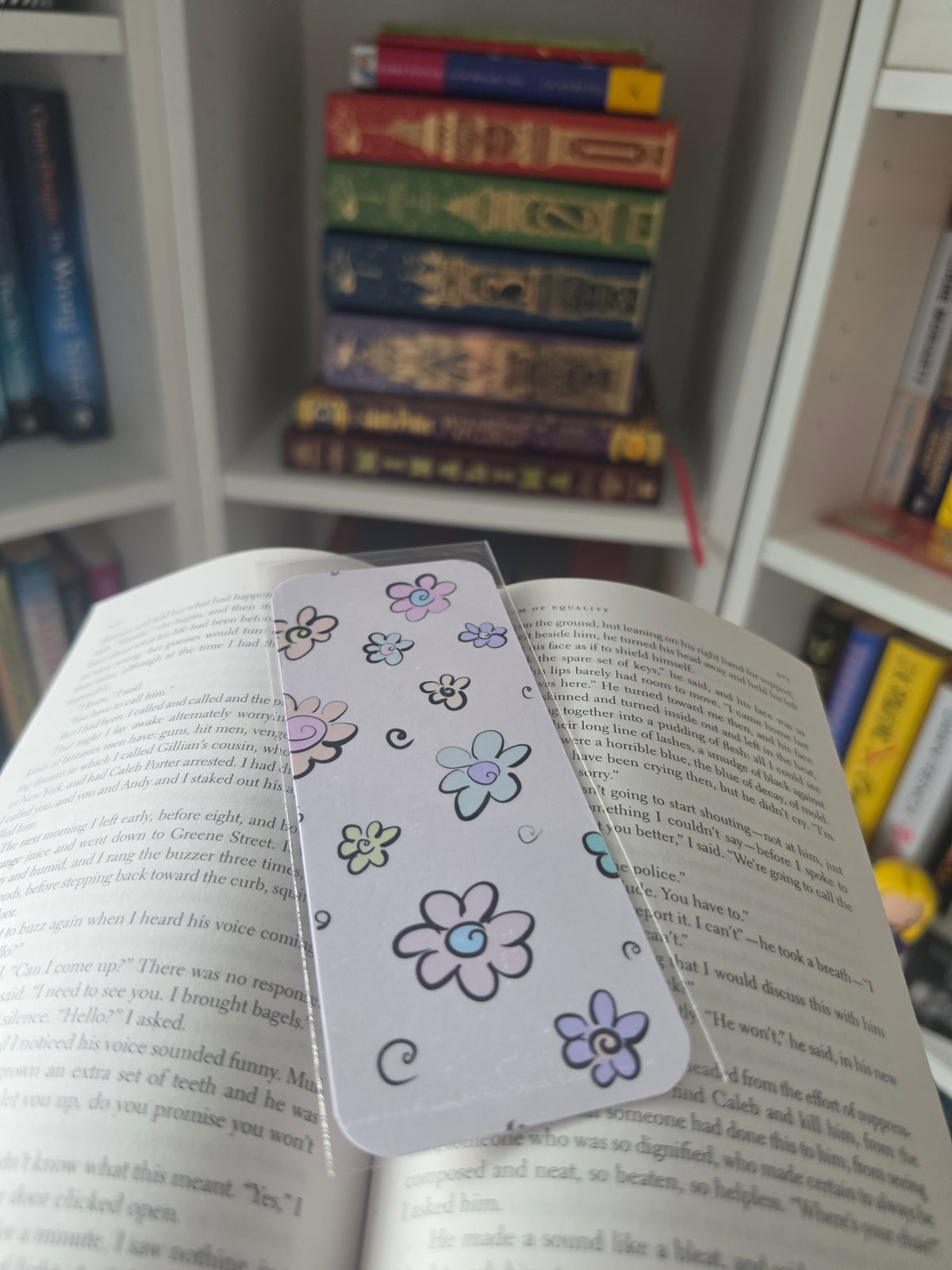 A white paper bookmark with colorful hand-drawn flowers and small swirls, resting on the pages of an open book in front of a blurred background of stacked books.