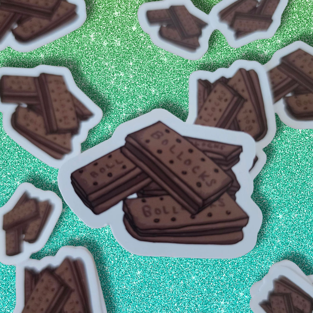 A pile of die-cut stickers featuring a well known brown chocolate biscuit treats with the word 'BOLLOCKS' printed on them, arranged on a bright glittery green background.