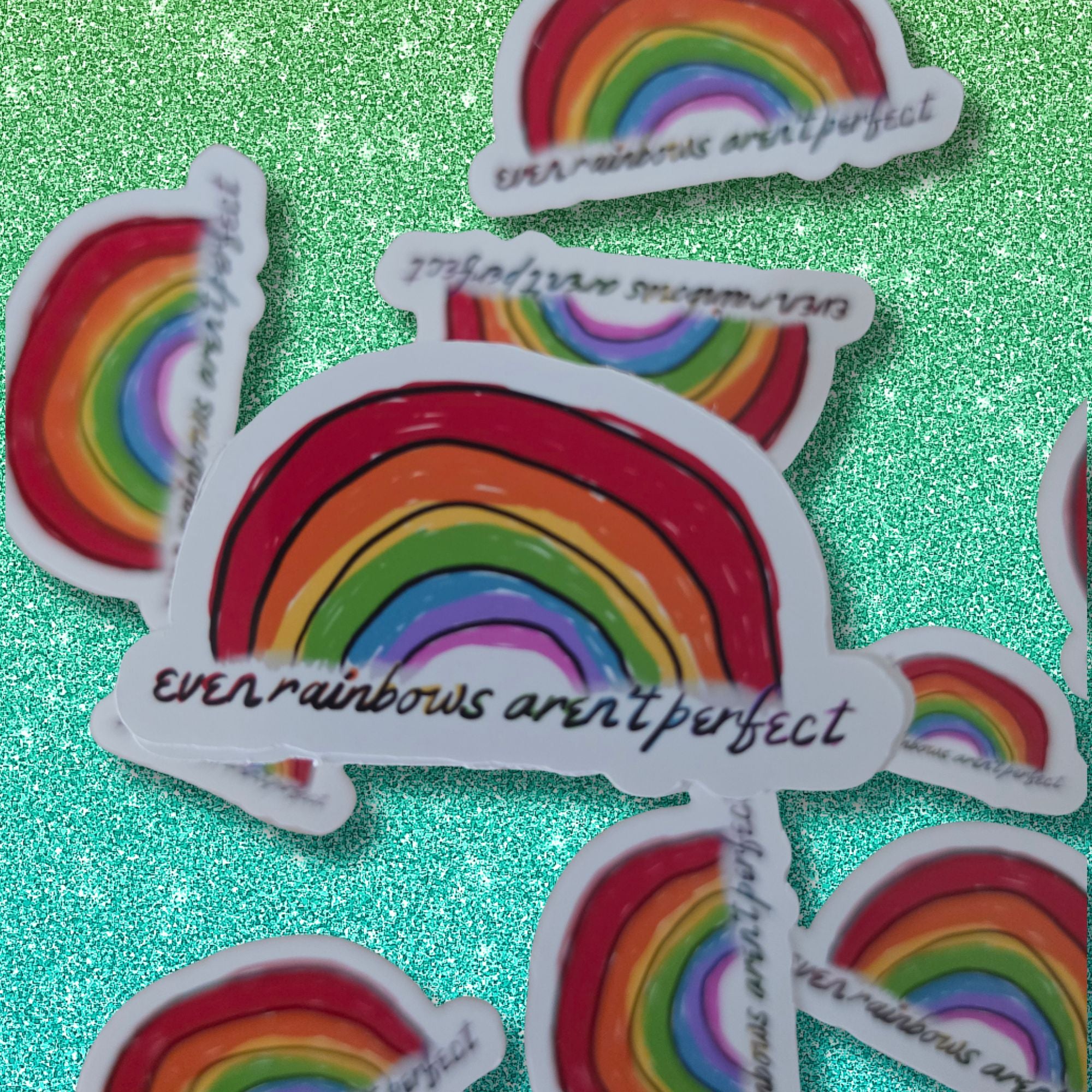 A set of die-cut stickers featuring a hand-drawn rainbow with the text 'even rainbows aren't perfect' in black script, arranged on a sparkling mint green background.