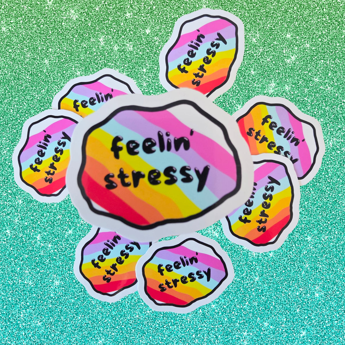 A set of die-cut stickers with a rainbow gradient background and the text 'feelin' stressy' in black script, arranged on a sparkling green background.
