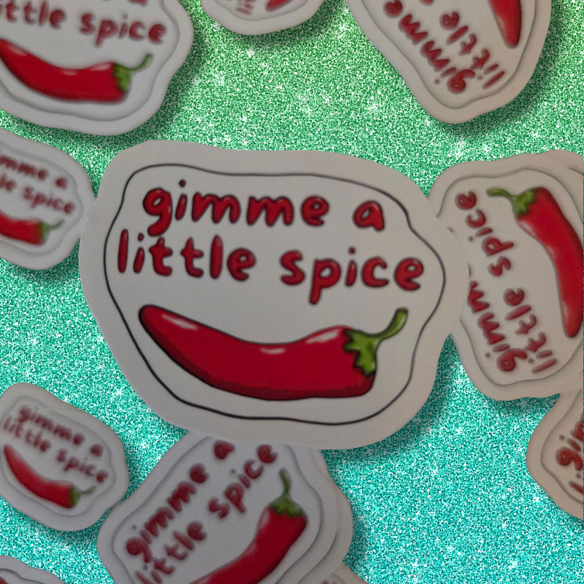 A close-up of multiple die-cut stickers featuring the phrase 'gimme a little spice' in red lettering above an illustration of a red chili pepper with green stem, arranged on a bright glittery aqua background.