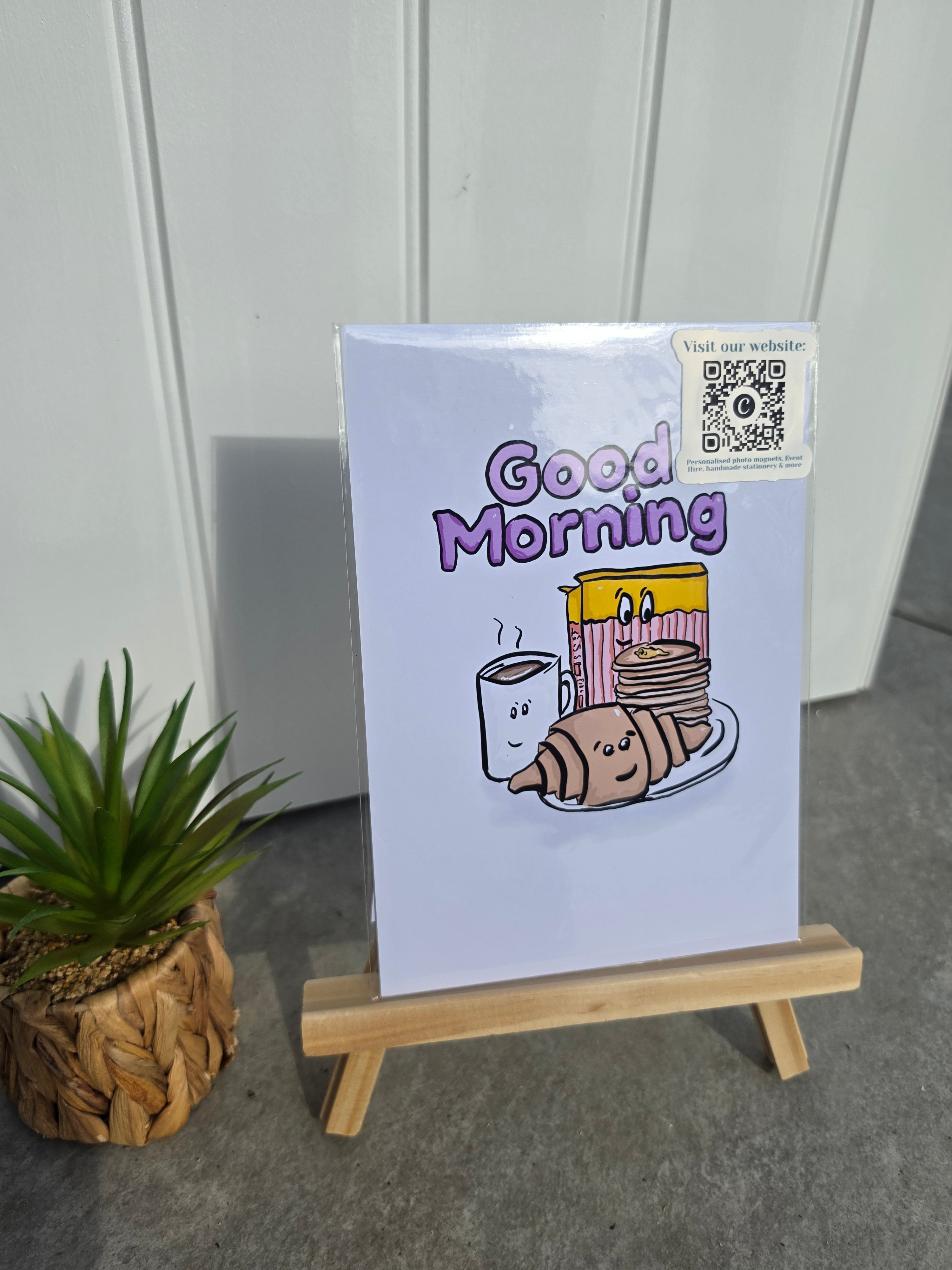 A square art print with a 'Good Morning' breakfast illustration displayed on a small wooden easel. The print features a light lavender background with purple text and a cartoon breakfast scene.