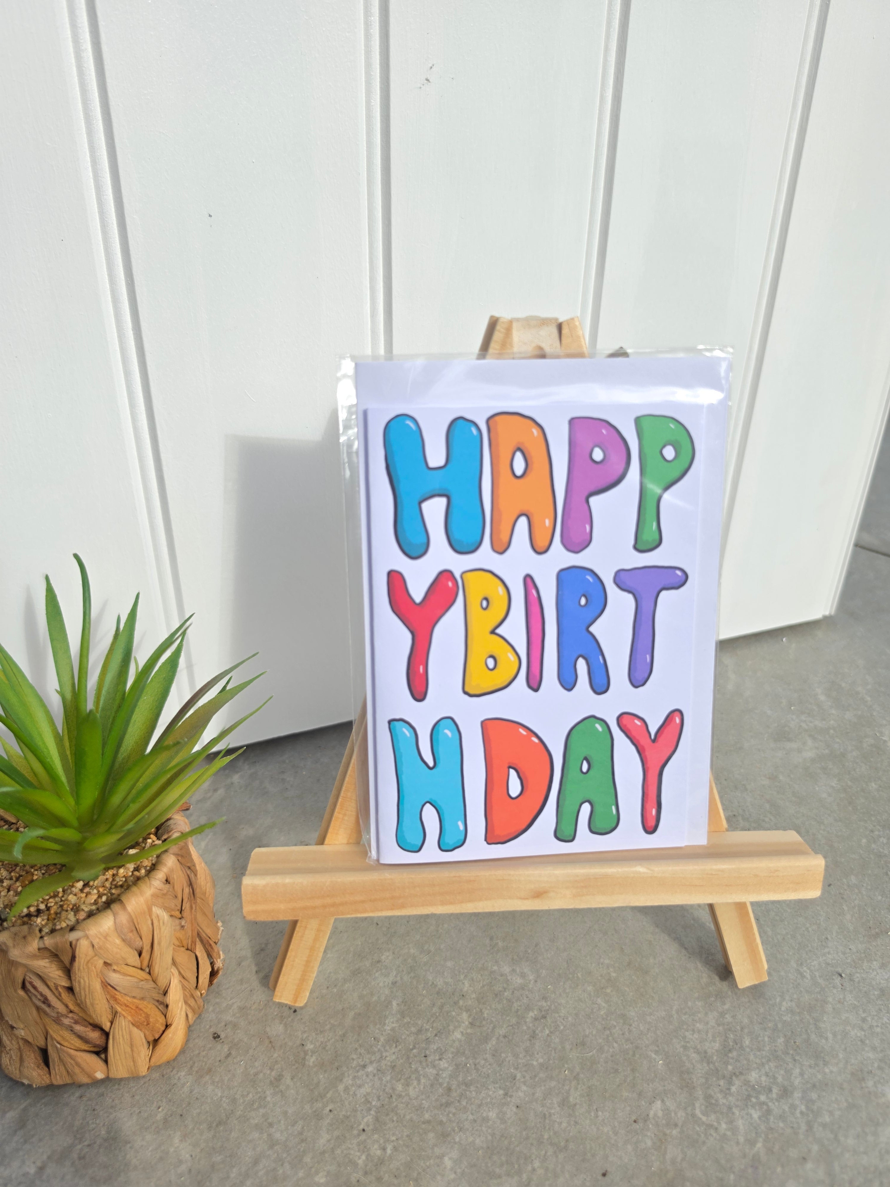A white card with colorful bubble letters spelling 'HAPPY BIRTHDAY' stands on a small wooden easel. The card is photographed indoors against a white paneled wall, with a potted succulent plant at the lower left.