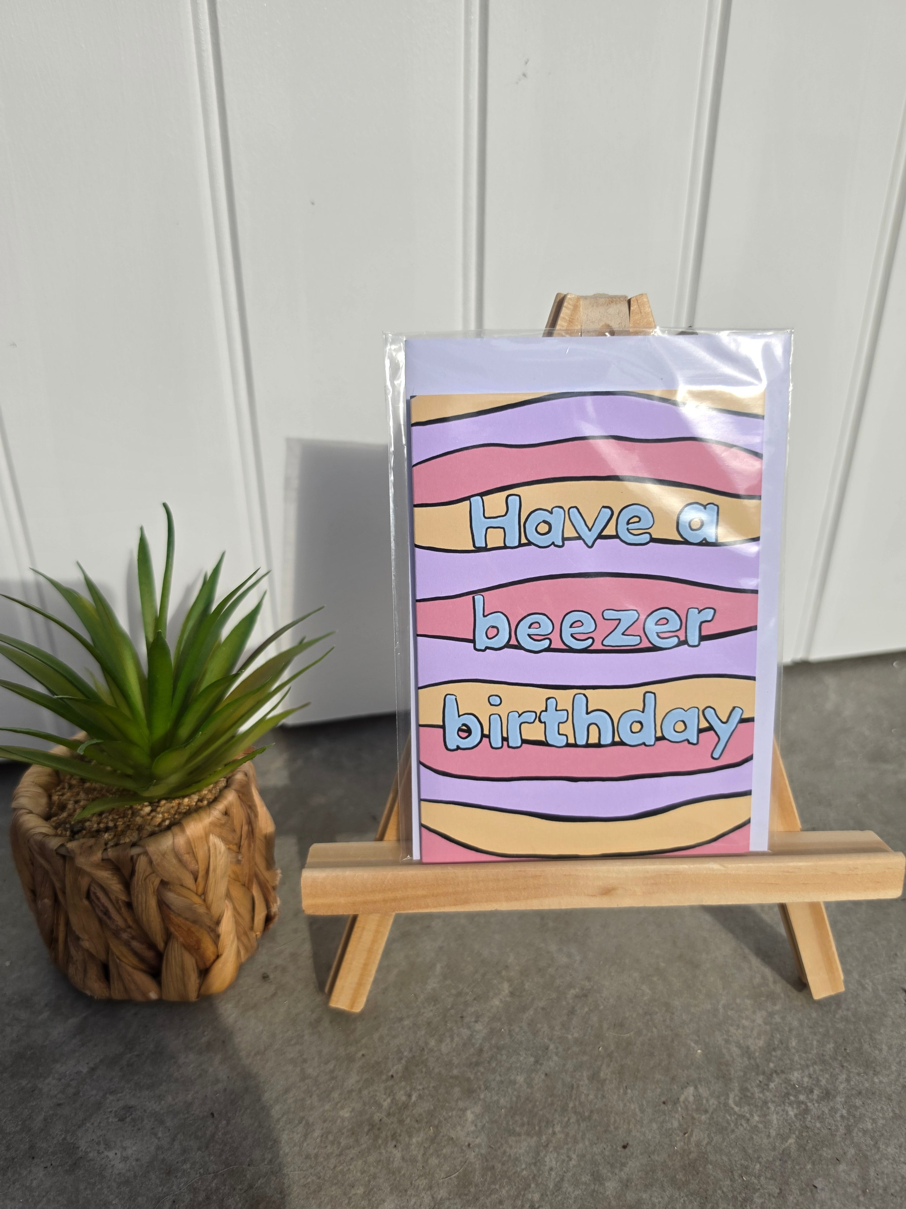 A greeting card with a pastel striped pattern and the text 'Have a beezer birthday' displayed on a small wooden easel, next to a potted succulent.