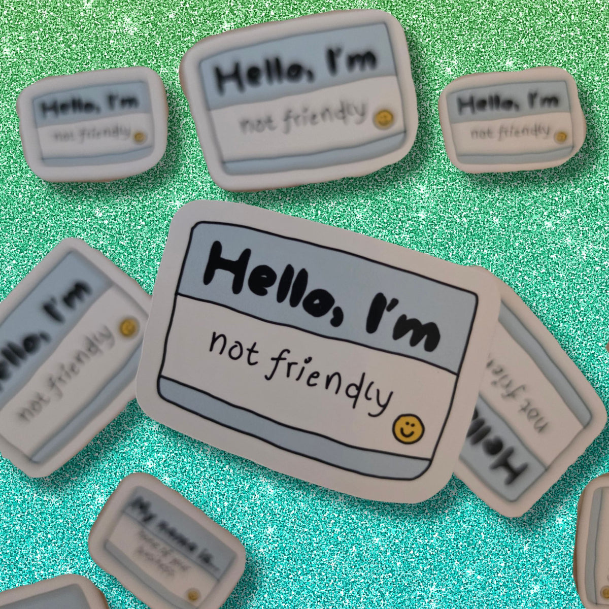 A set of cute, cartoon-style name tag-shaped stickers with the text 'Hello, I'm not friendly' and a small sad smiley face, arranged on a sparkling green glitter background.