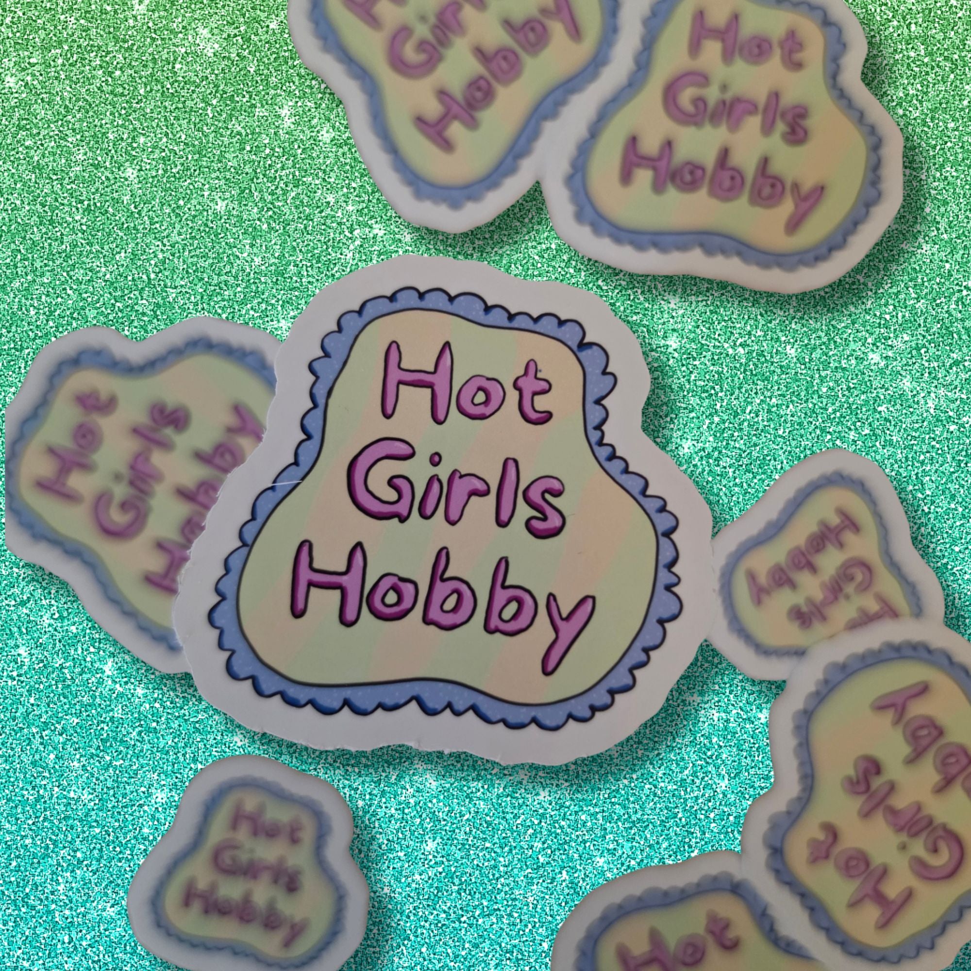 A set of die-cut stickers featuring the text 'Hot Girls Hobby' in purple lettering on a pale yellow background, outlined with a lavender-blue scalloped border. The stickers are arranged on a sparkling teal surface.