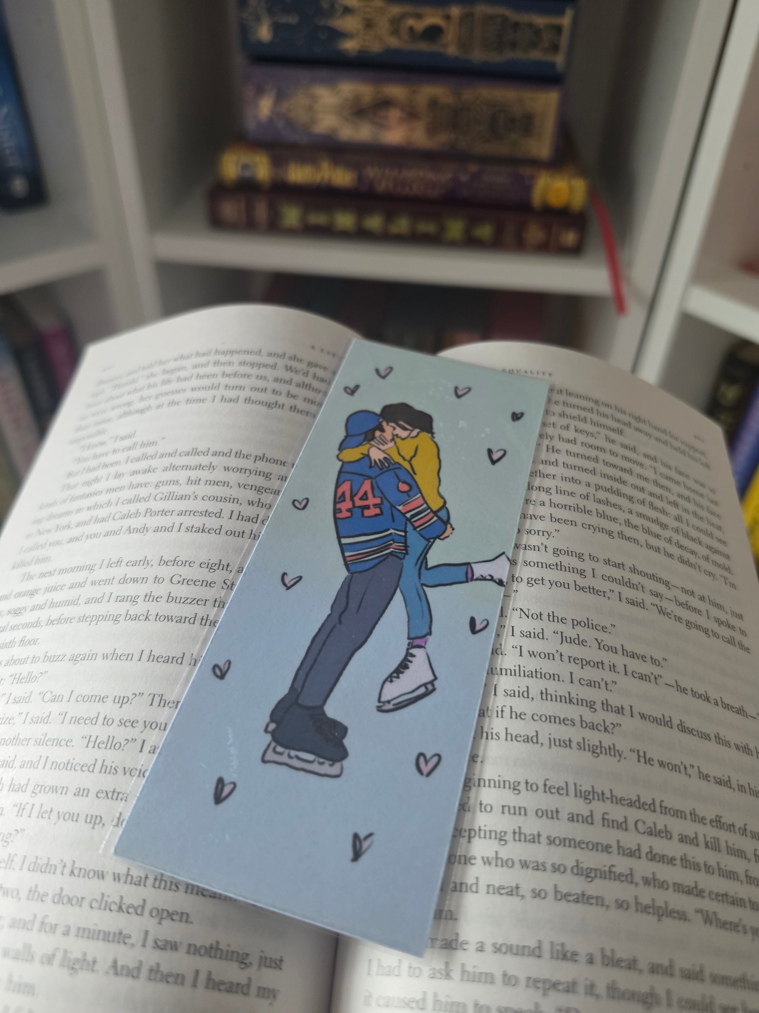 A light blue bookmark with a cartoon illustration of two ice skaters and small heart doodles, placed on open book pages with other books blurred in the background.