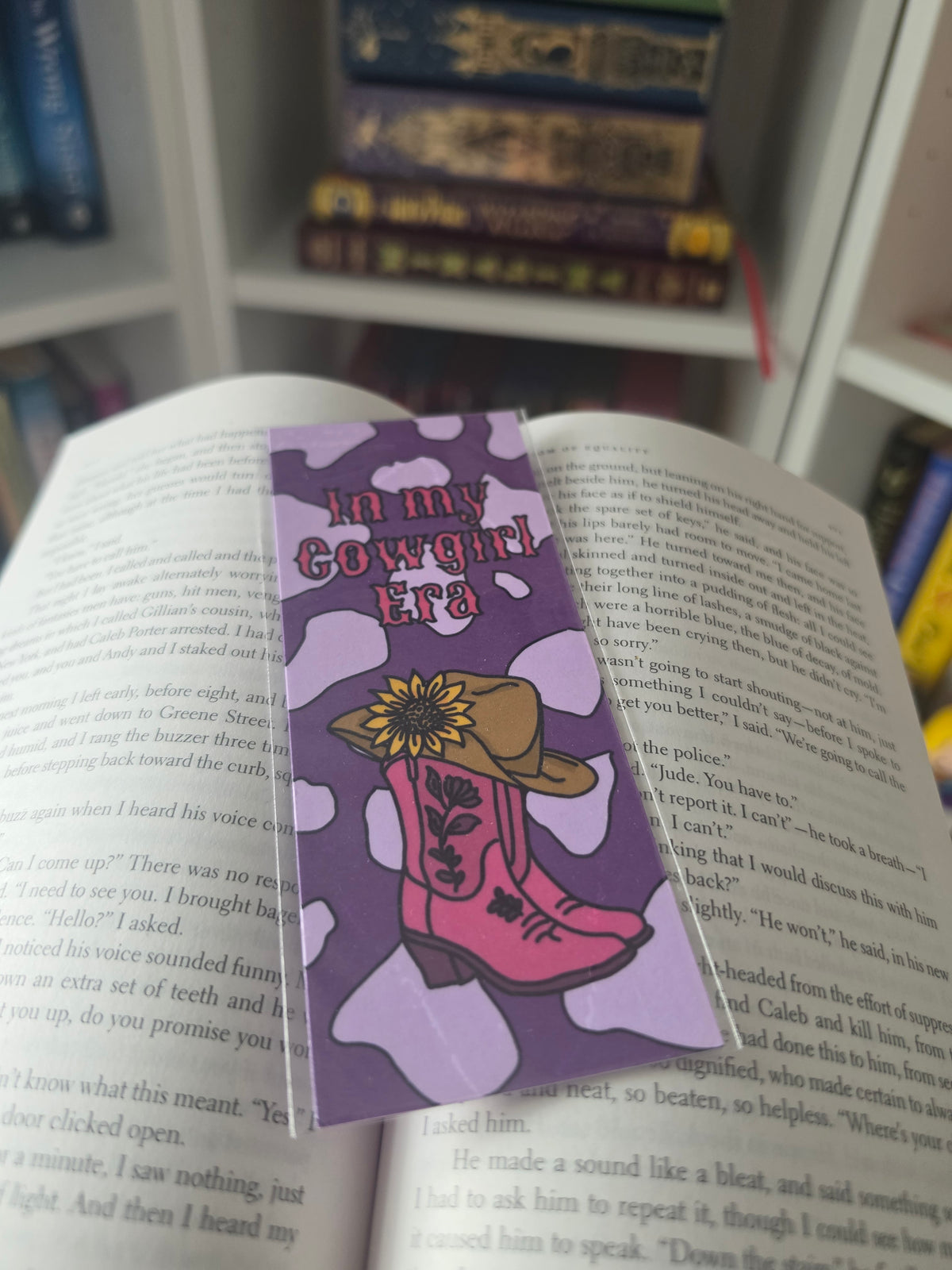 A purple book bookmark with the text 'In my cowgirl era' and an illustration of pink cowboy boots, a yellow flower, and a brown hat, placed on an open book with other books blurred in the background.