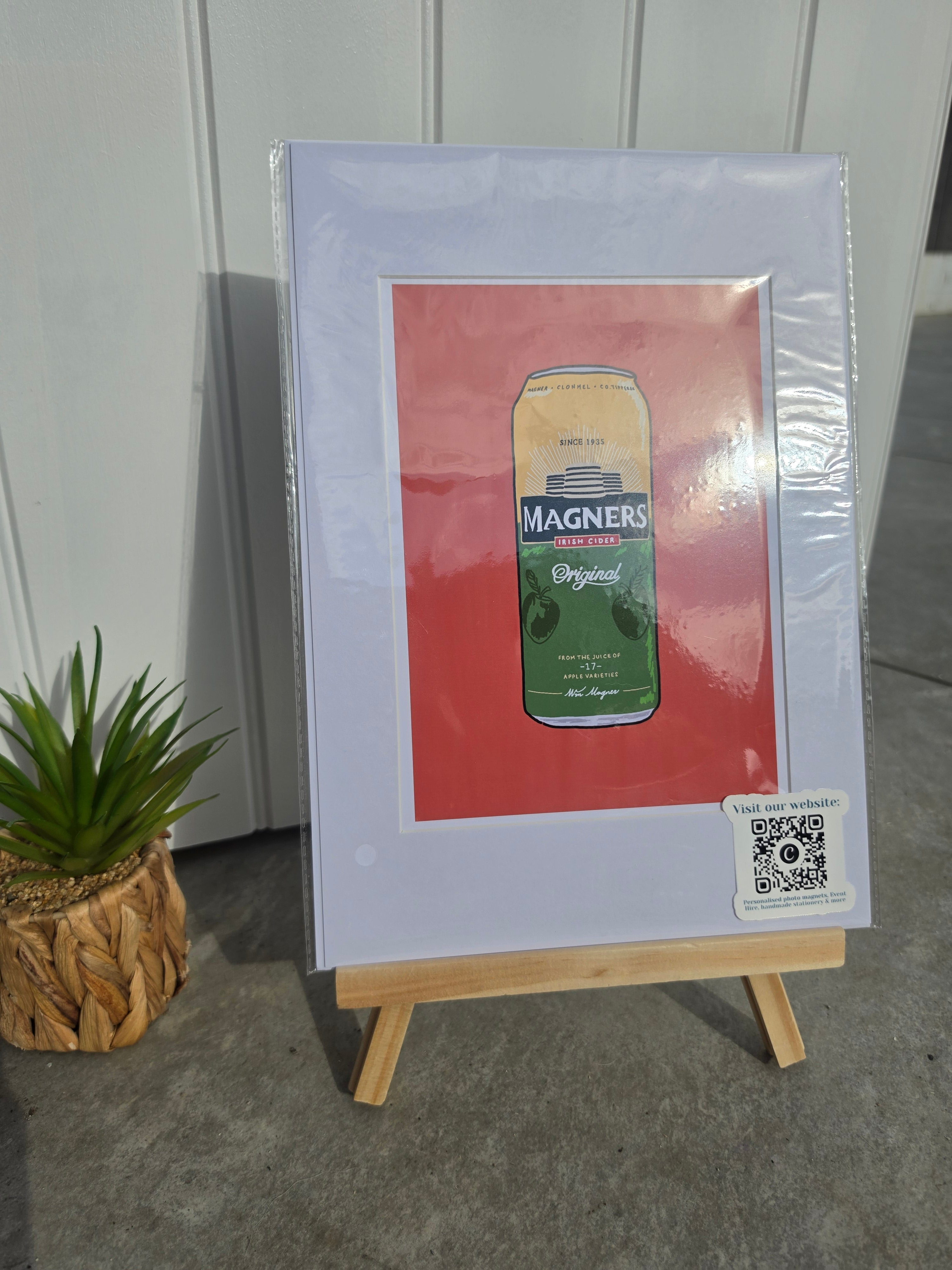 A framed art print featuring a vintage-style Magners beer can illustration on a red background, displayed on a small wooden easel. 