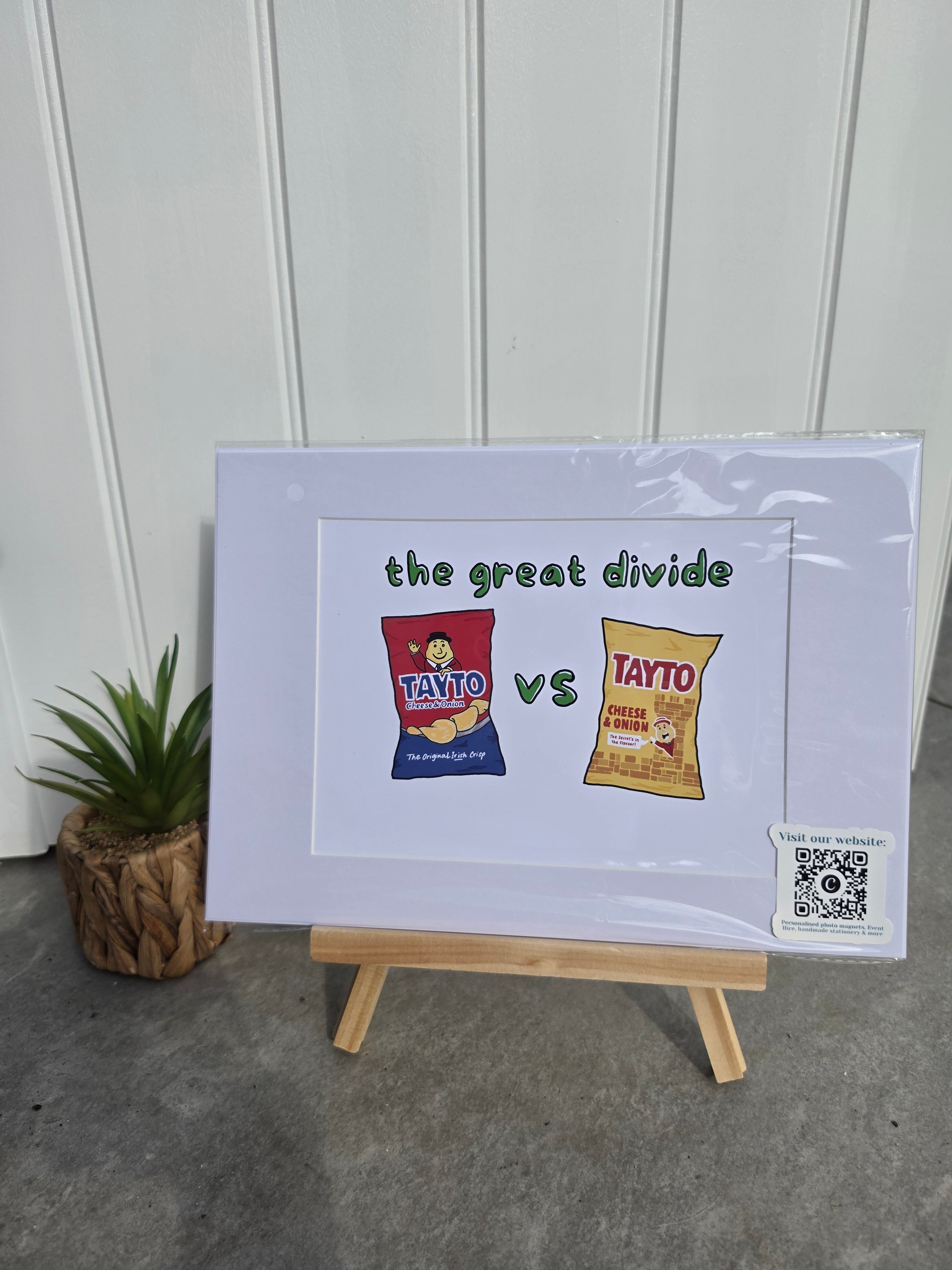 A framed print on a small wooden easel, featuring a comparison graphic between a red and a yellow crisp bag with the text 'the great divide vs'. The print is wrapped in protective plastic, set against a white paneled wall with a small potted plant on the left.