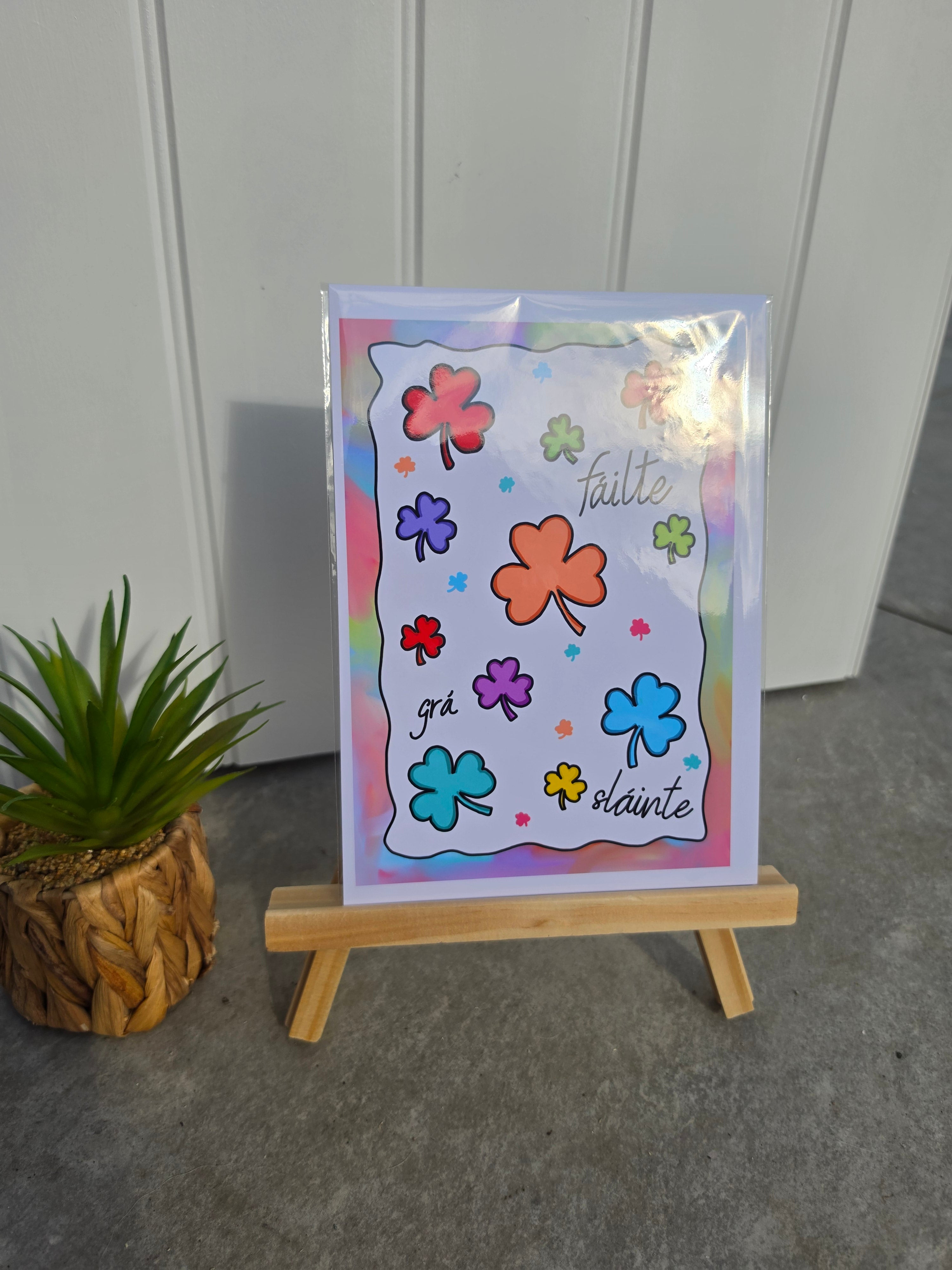 A square printed card or art print with colorful shamrock flowers and the words 'taille grâ' and 'slainte', displayed upright on a small wooden easel. A potted green plant sits to the left, and a light paneled wall and gray floor are in the background.