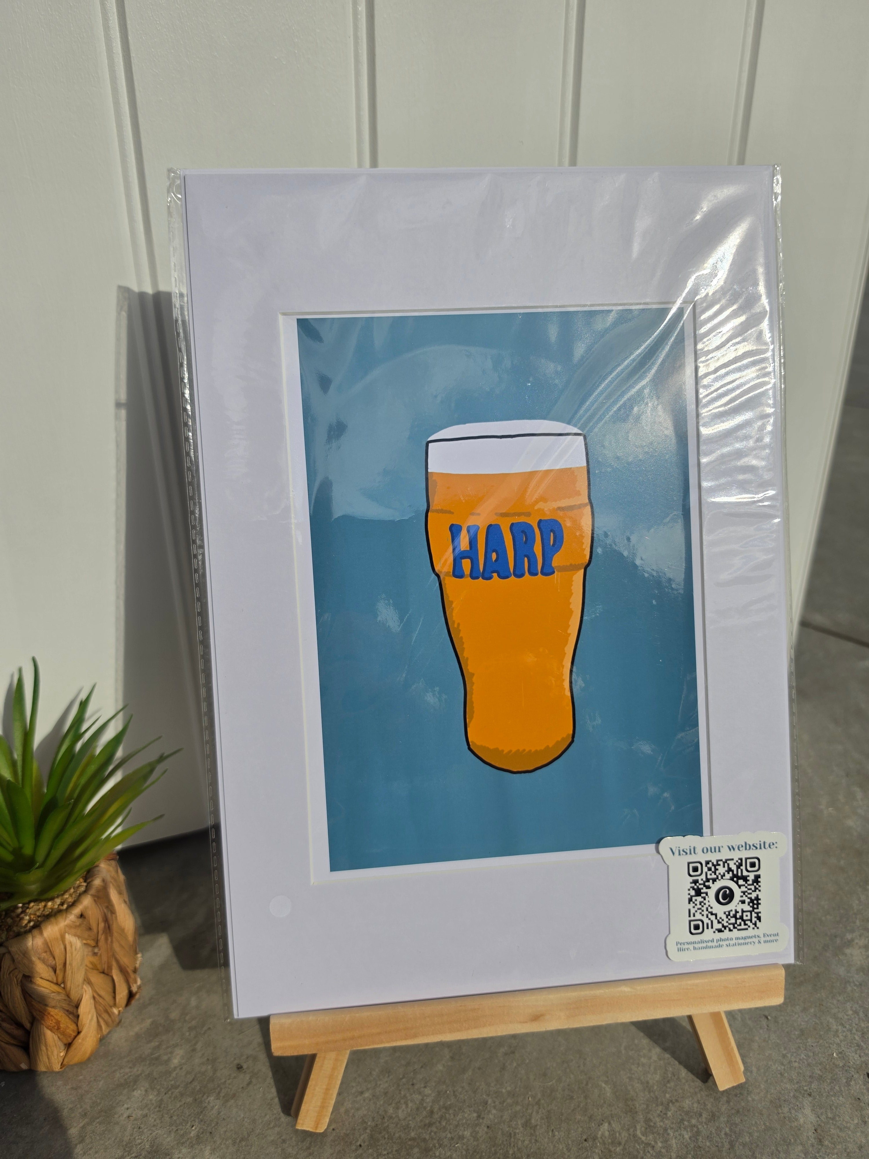 A framed art print on a small wooden easel, featuring a stylized orange beer glass with blue 'HARP' lettering against a blue background. The print is inside a white mat and clear protective plastic, photographed outdoors against a white paneled wall with a small potted plant at the side.