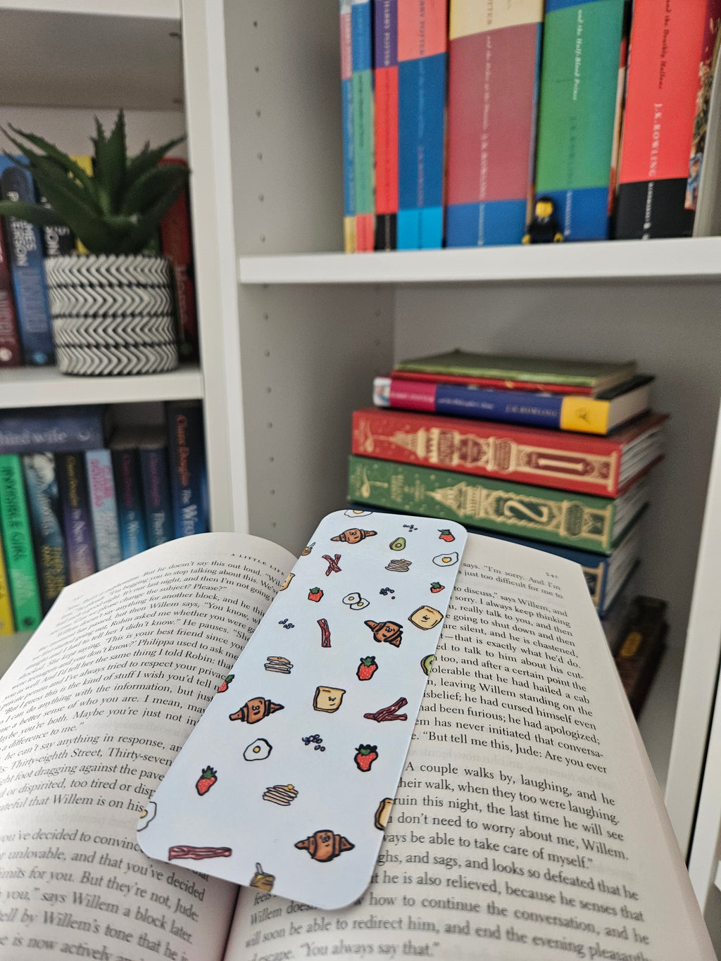 A bookmark with colorful cartoon food illustrations rests across open book pages, with a bookshelf and stacked books in the background.