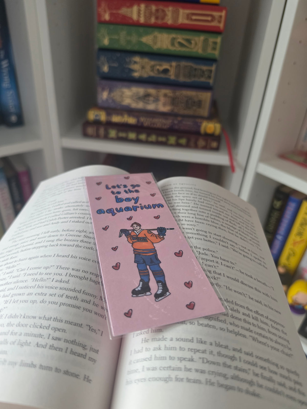 A pink paper bookmark with a cartoon boy holding a hockey stick and the text 'Let's go to the boy aquarium' is placed on an open book, with shelves of books blurred in the background.