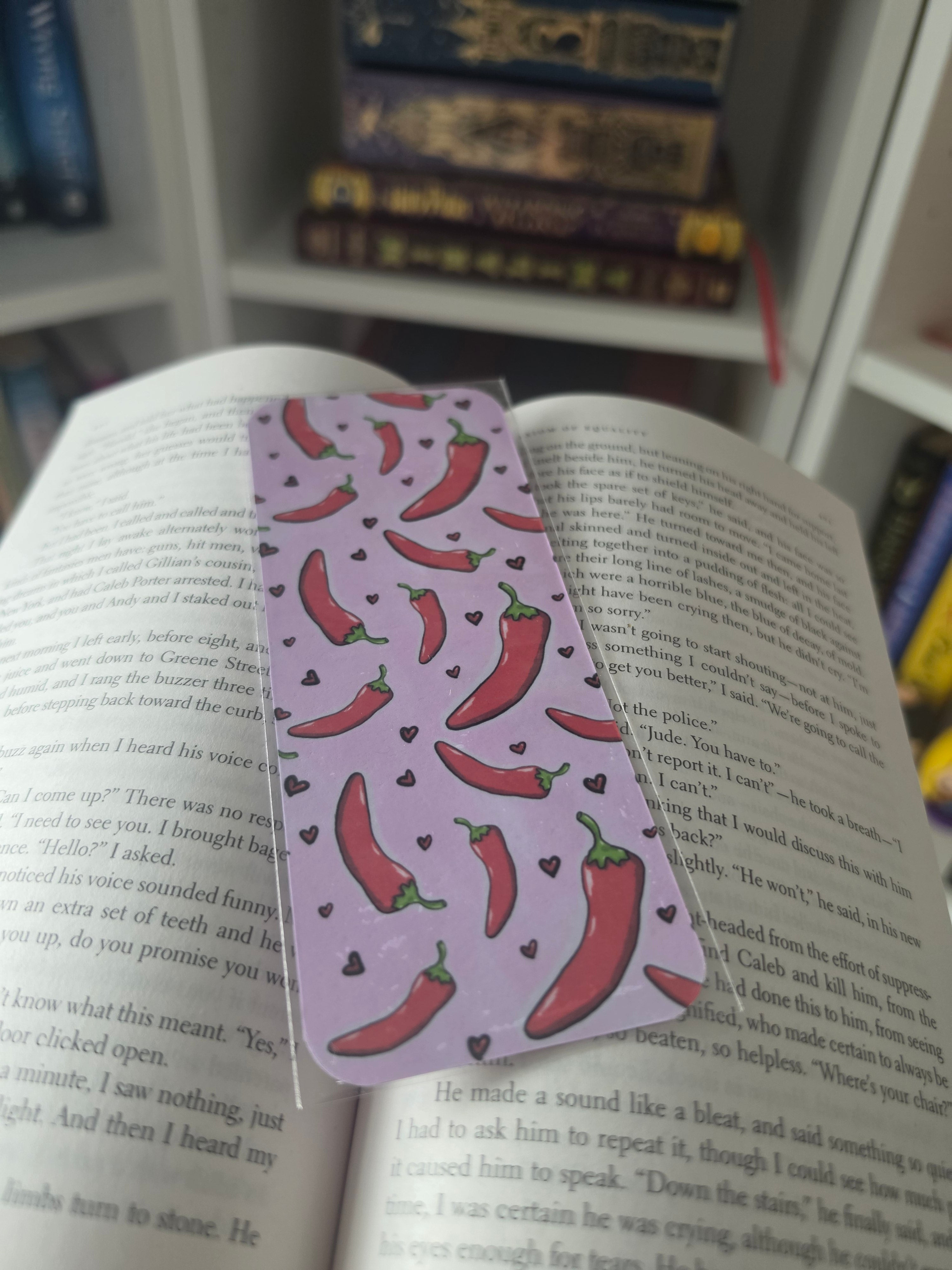 A lavender bookmark featuring a repeating pattern of red chili peppers and small black hearts, resting on an open book with other books blurred in the background.