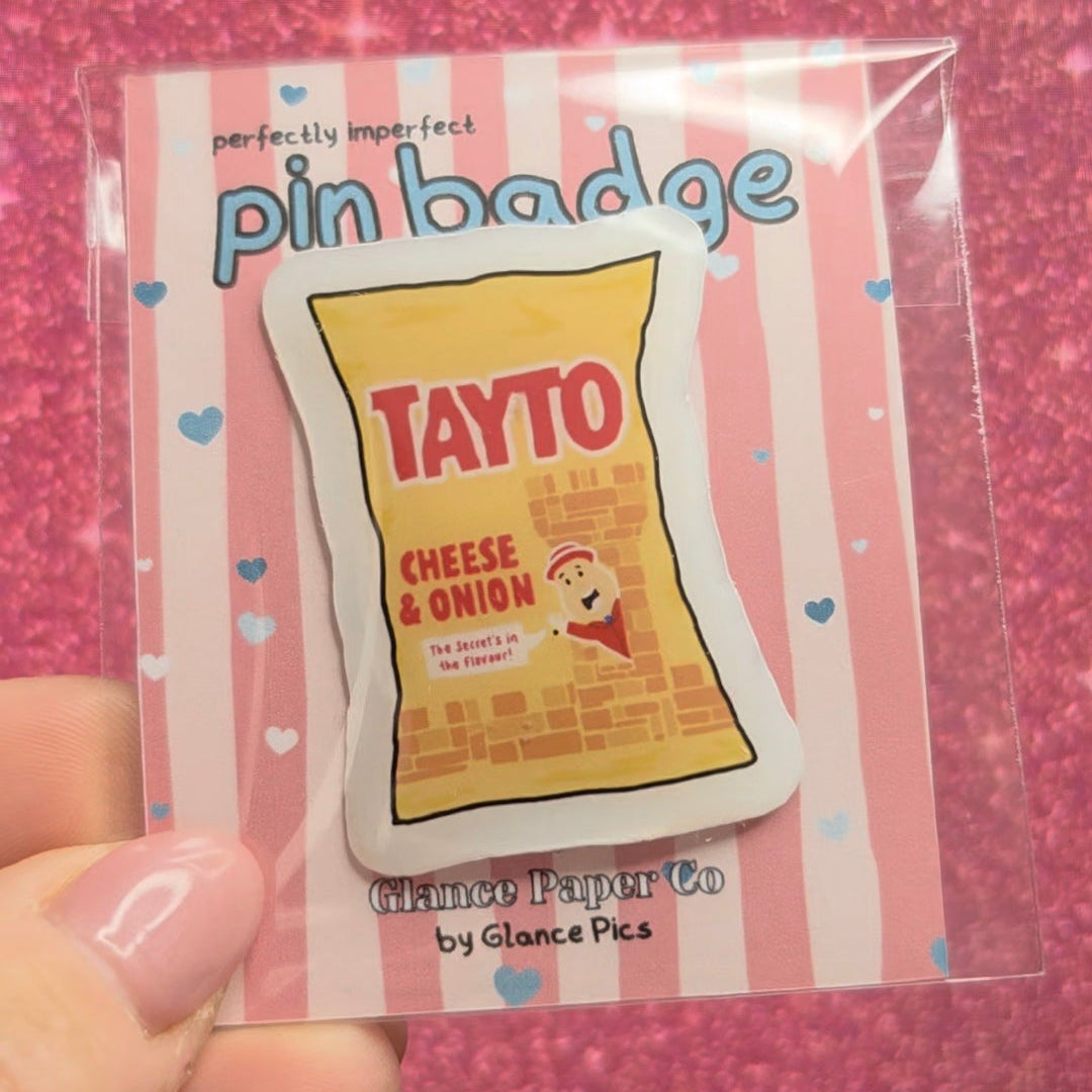 A hand holding a clear plastic sleeve containing a pin badge shaped like a bag of Tayto Cheese & Onion chips, set against a sparkly pink background.