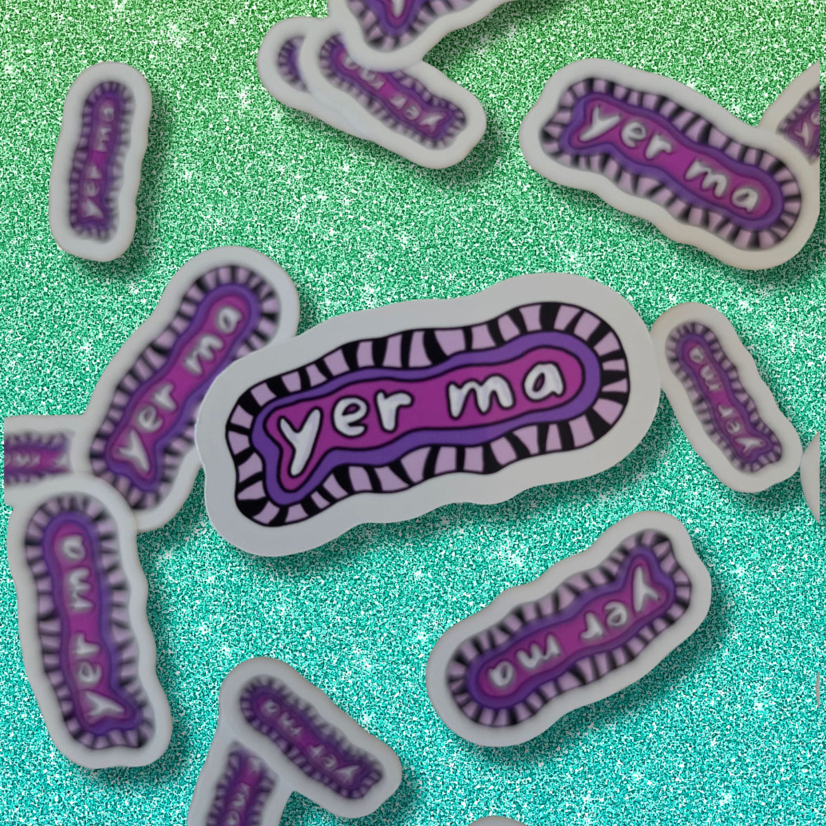 Several small die-cut stickers shaped like a stylized bacterium are scattered on a glittery turquoise background. The stickers have a white border, a purple center, black striped edges, and the word "yer ma" in white lettering.