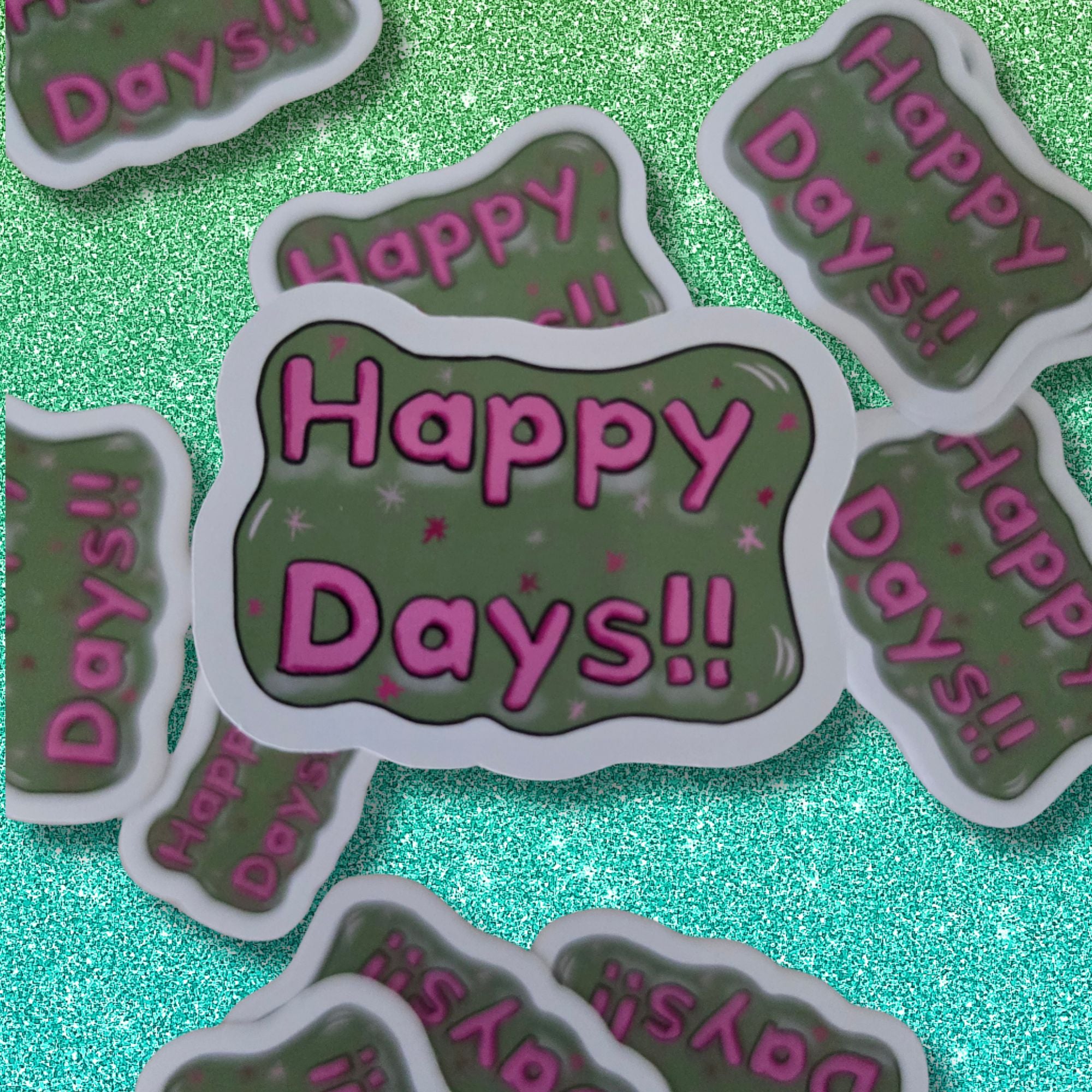 A close-up of several matching decorative stickers or patches scattered on a glittery turquoise background. The central piece shows the words "Happy Days!!" in pink lettering on a muted green shape with a white border.