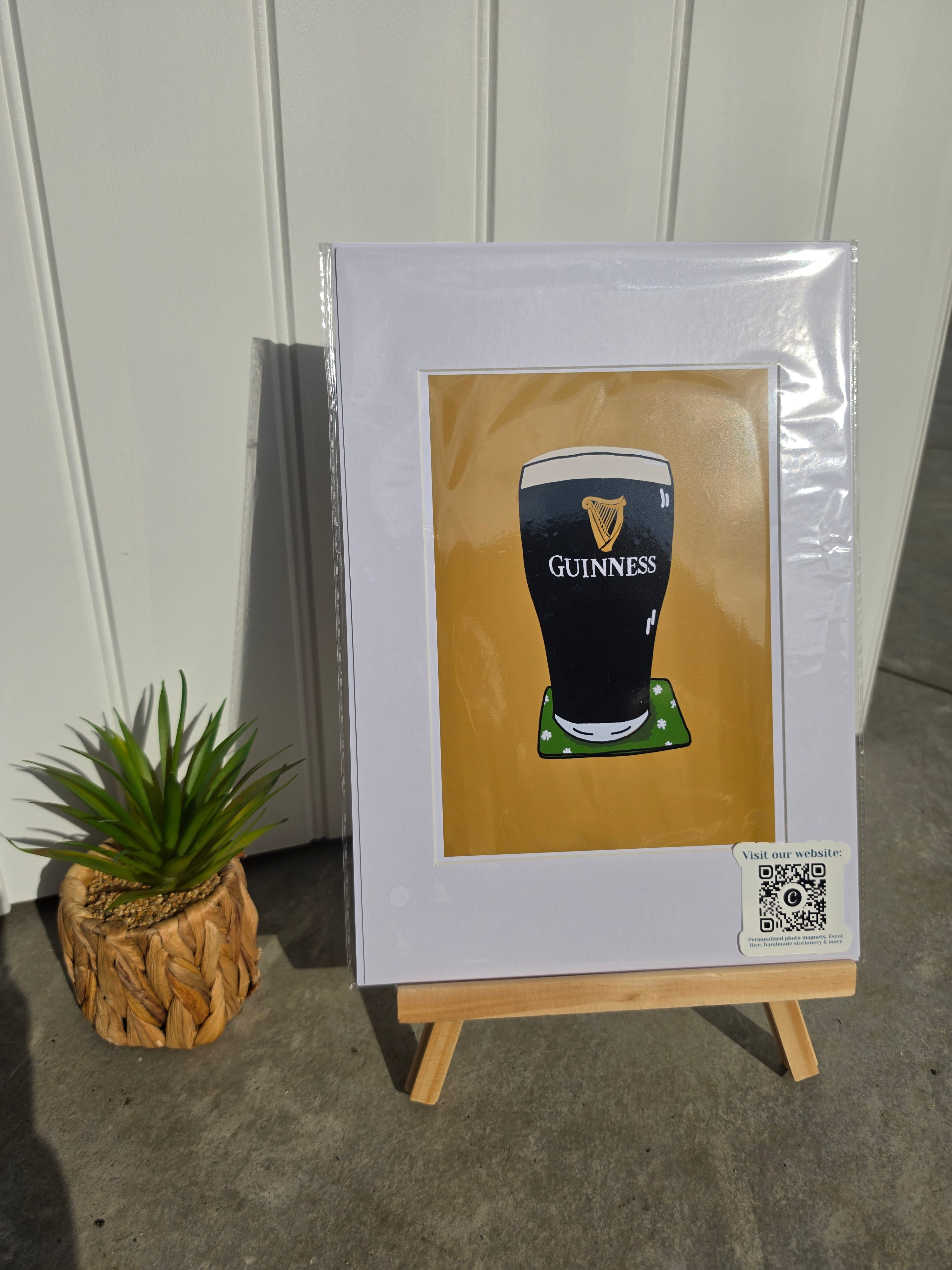 A framed art print featuring a pint of Guinness beer, displayed on a small wooden easel next to a small potted succulent plant. The print has a white mat border and a gold background