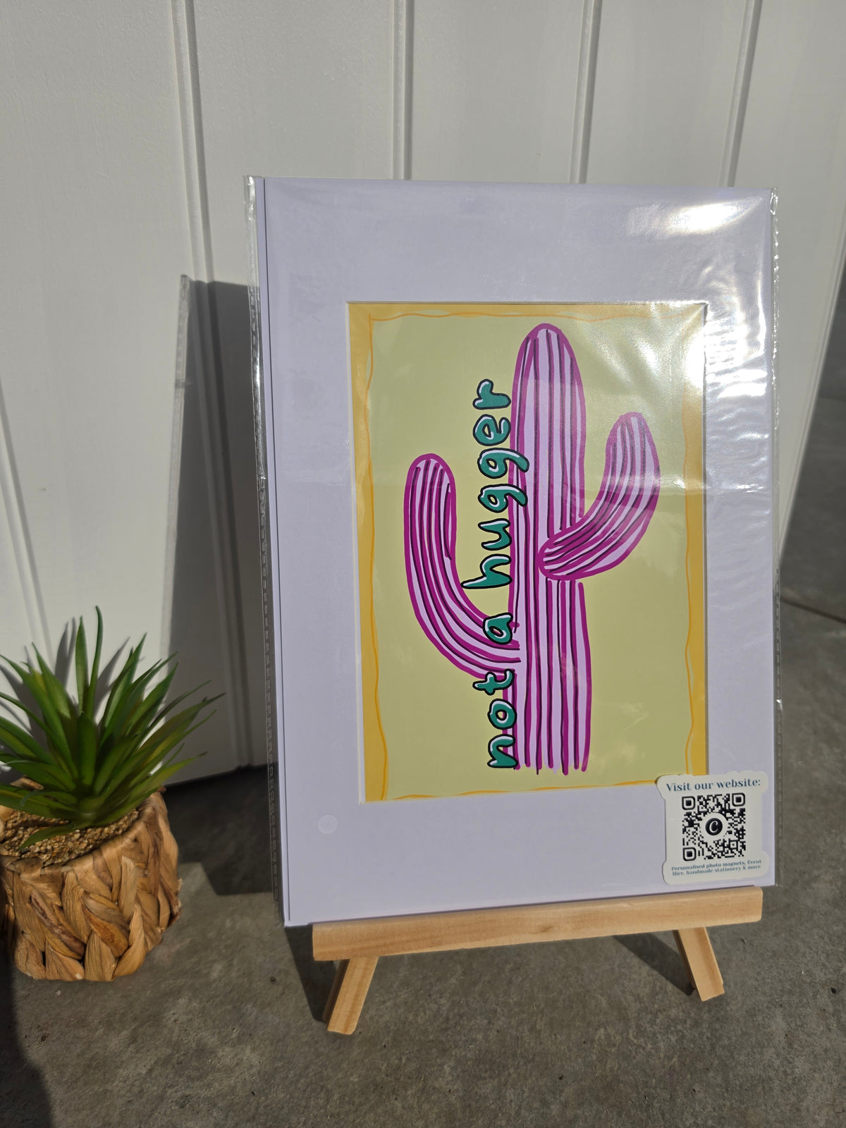 A framed art print displayed on a small wooden easel, featuring a stylized purple cactus with the text 'not a hugger' on a pale yellow background. The print is wrapped in protective plastic, and a small potted succulent sits to the left against a white paneled wall.
