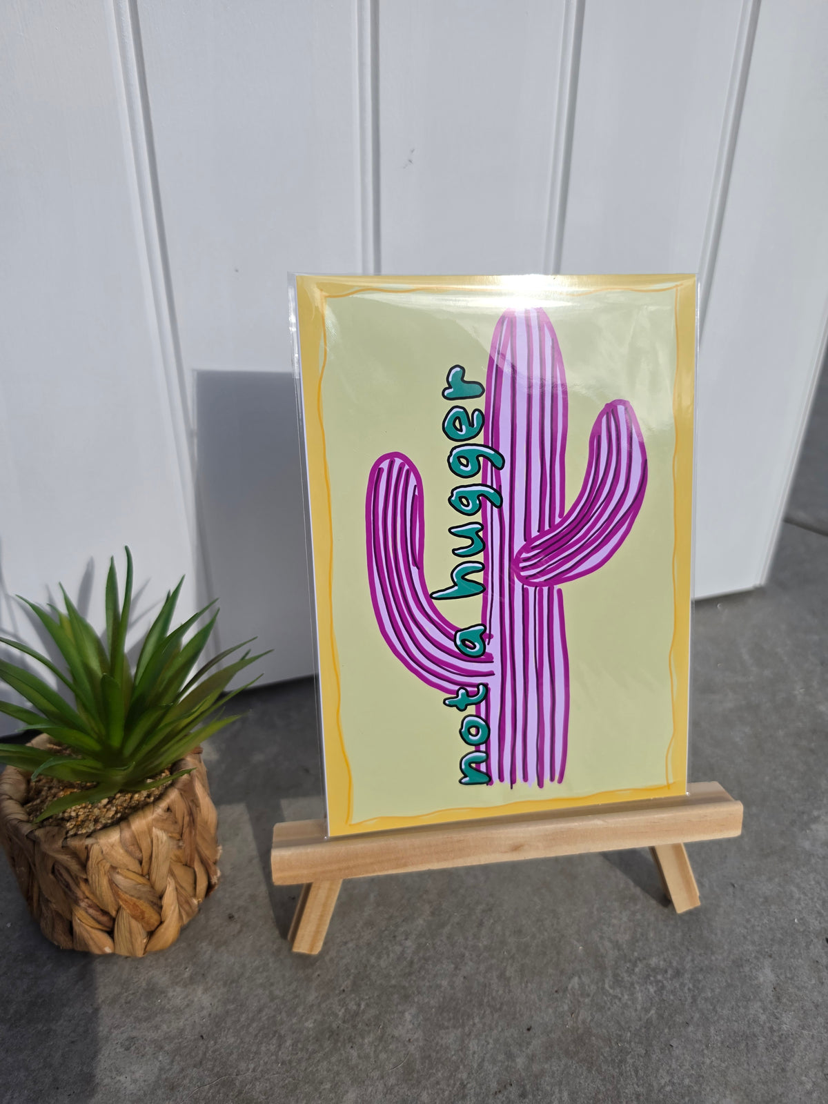 A square art print featuring a purple striped cactus and the text 'not a hugger' in teal, displayed on a small wooden easel next to a potted succulent against a white paneled wall.