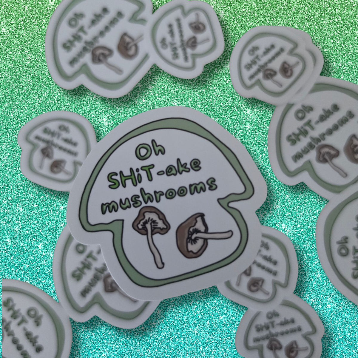 A close-up of several die-cut stickers featuring a mushroom illustration and the text 'Oh SHiT-ake mushrooms' arranged on a glittery green background.