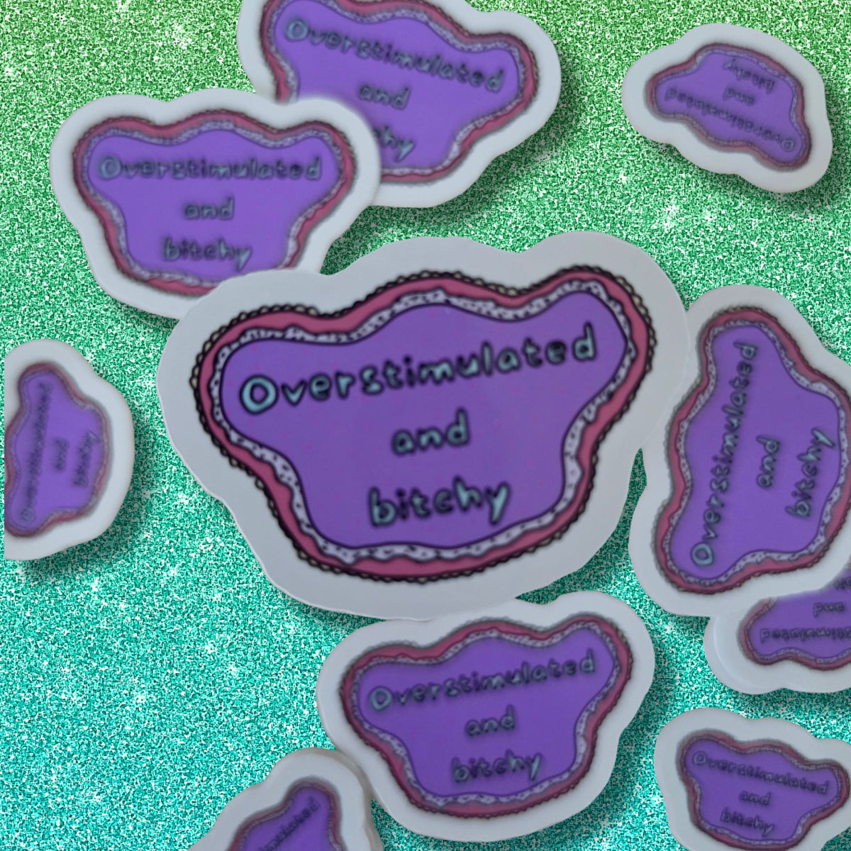 A set of die-cut stickers shaped like a purple swirl with the text 'Overstimulated and bitchy' in white lettering, displayed on a sparkling green background.